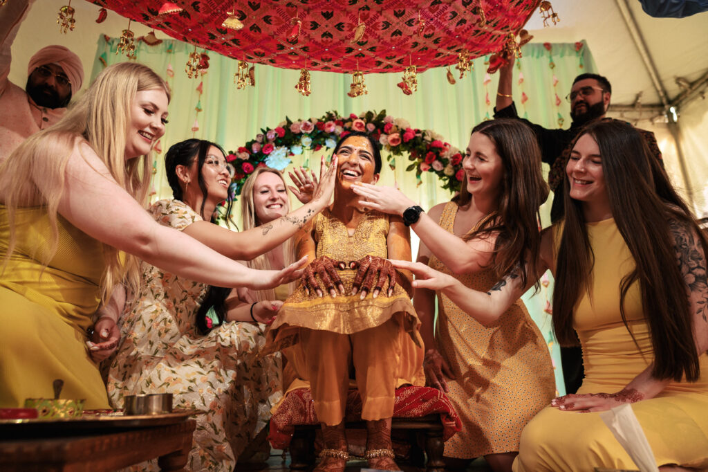 A joyful woman in traditional yellow attire sits at the center while friends, also dressed in yellow and white outfits, gather around her with hands extended playfully. The backdrop includes vibrant decorations and floral arrangements, creating a festive atmosphere.