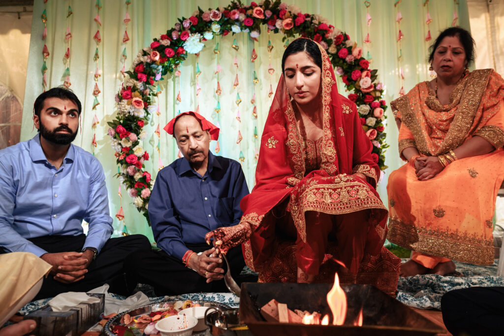 A woman in a red traditional outfit participates in a ceremonial ritual involving fire, flanked by two men and another woman on a decorated stage with a floral backdrop. The group appears solemn and focused on the ritual.