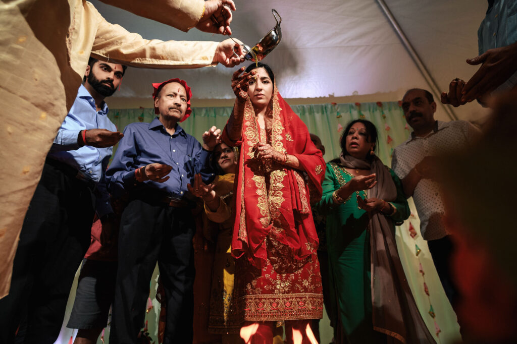 A woman wearing a red traditional outfit participates in a ceremonial event under a tent with several other people surrounding her. One person pours a liquid from a small vessel into her hand. The group appears engaged in the ritual.