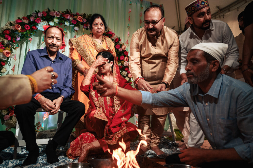 A group of people participate in a traditional ceremony. In the center, a woman in red covers her face. A man in blue and a woman in a yellow saree stand behind, while others gather around, some performing rituals near a fire. Floral decorations adorn the background.