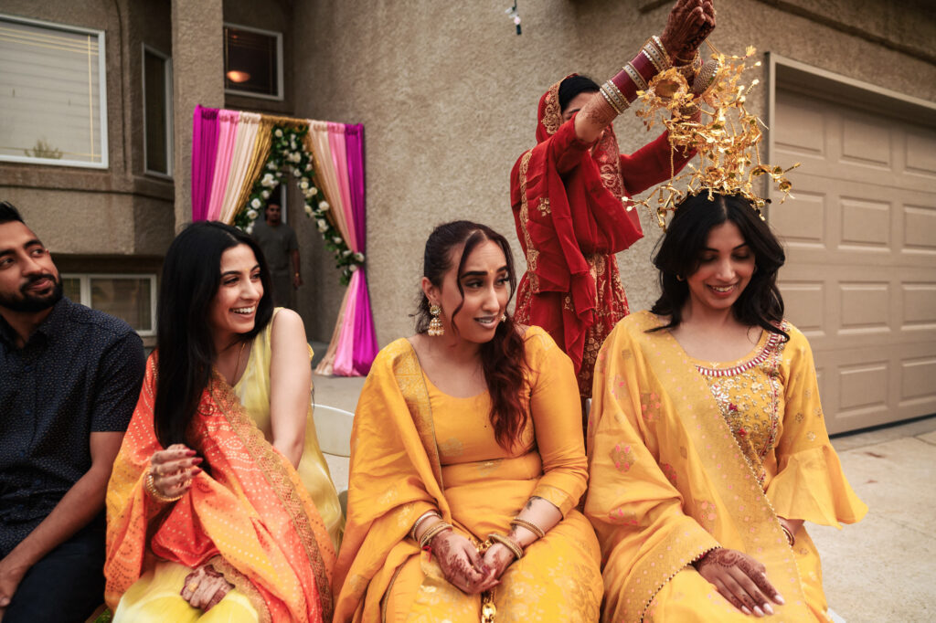 A group of people dressed in traditional Indian attire is sitting and smiling. A woman in a red outfit stands behind them, holding a decorative item above another woman's head, possibly as part of a ritual. The setting appears to be a home with festive decorations.