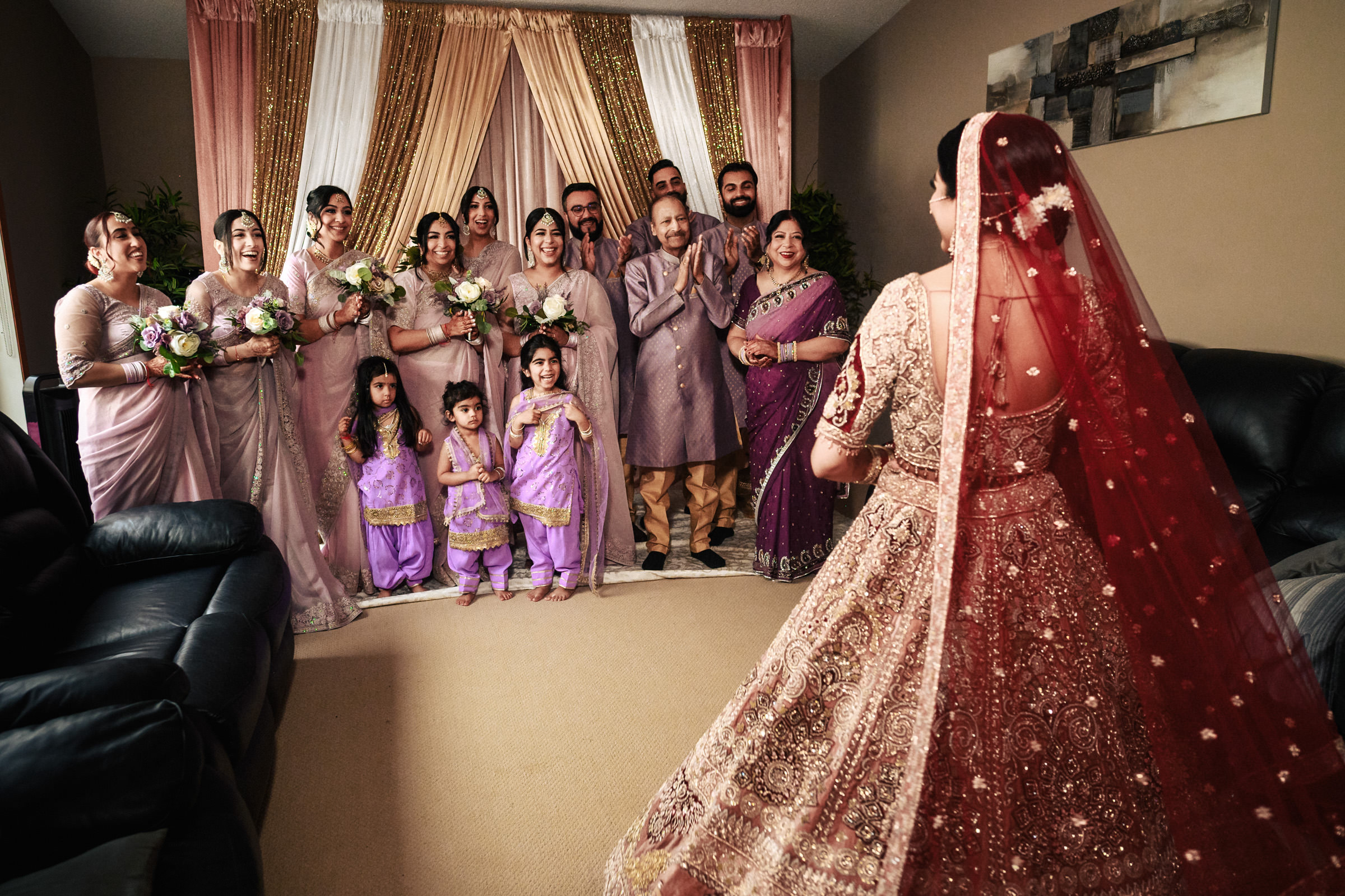 A bride in a traditional red wedding dress faces a group of family members and loved ones in a warmly lit room. They are dressed in coordinated purple and lavender outfits and appear joyful and celebratory as they look towards the bride.