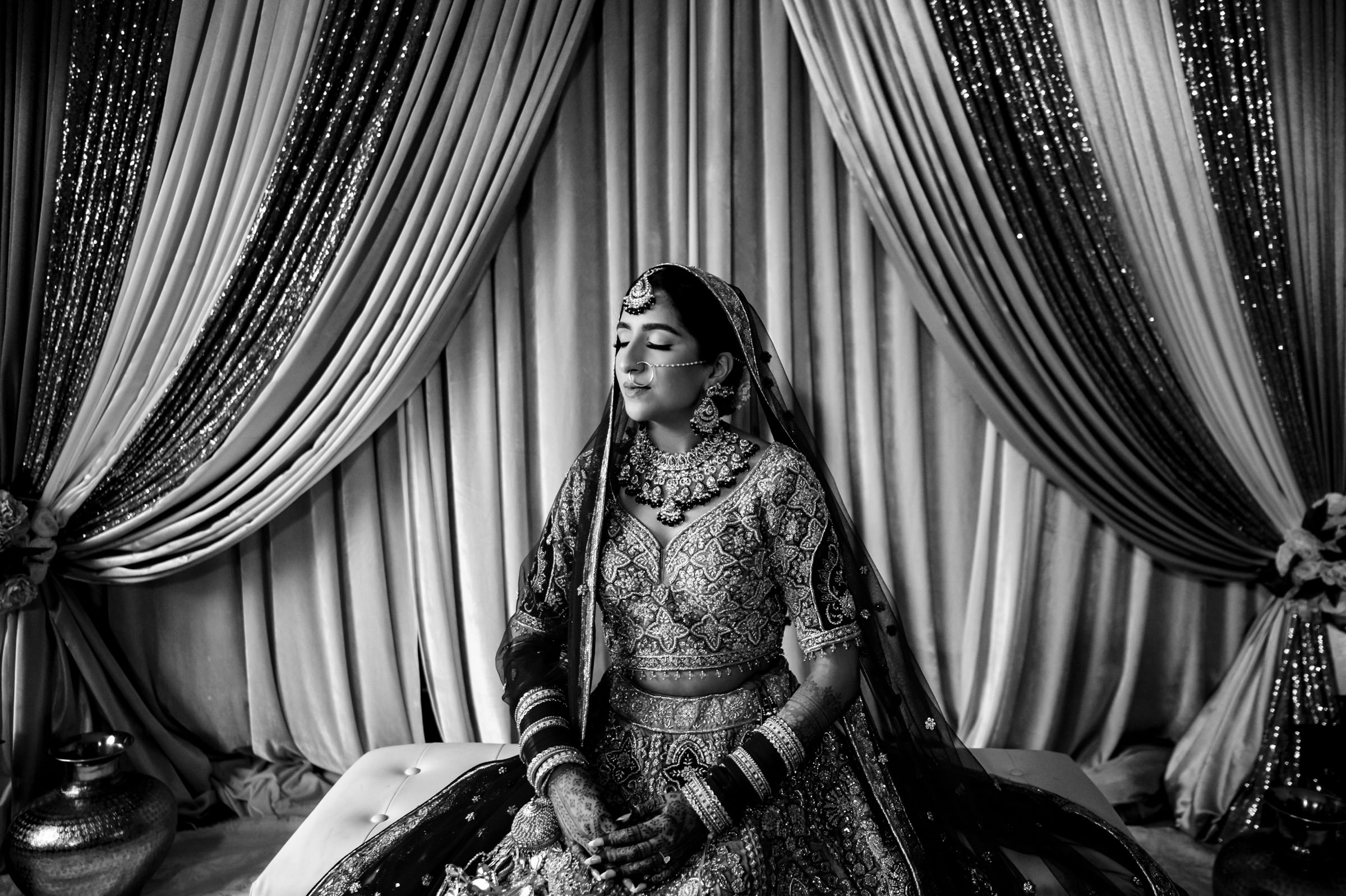 A bride sits with her eyes closed in front of a backdrop of draped, sequined curtains. She is adorned in traditional bridal attire, including a heavily embellished lehenga, ornate jewelry, bangles, and a headpiece, capturing a serene and contemplative moment.