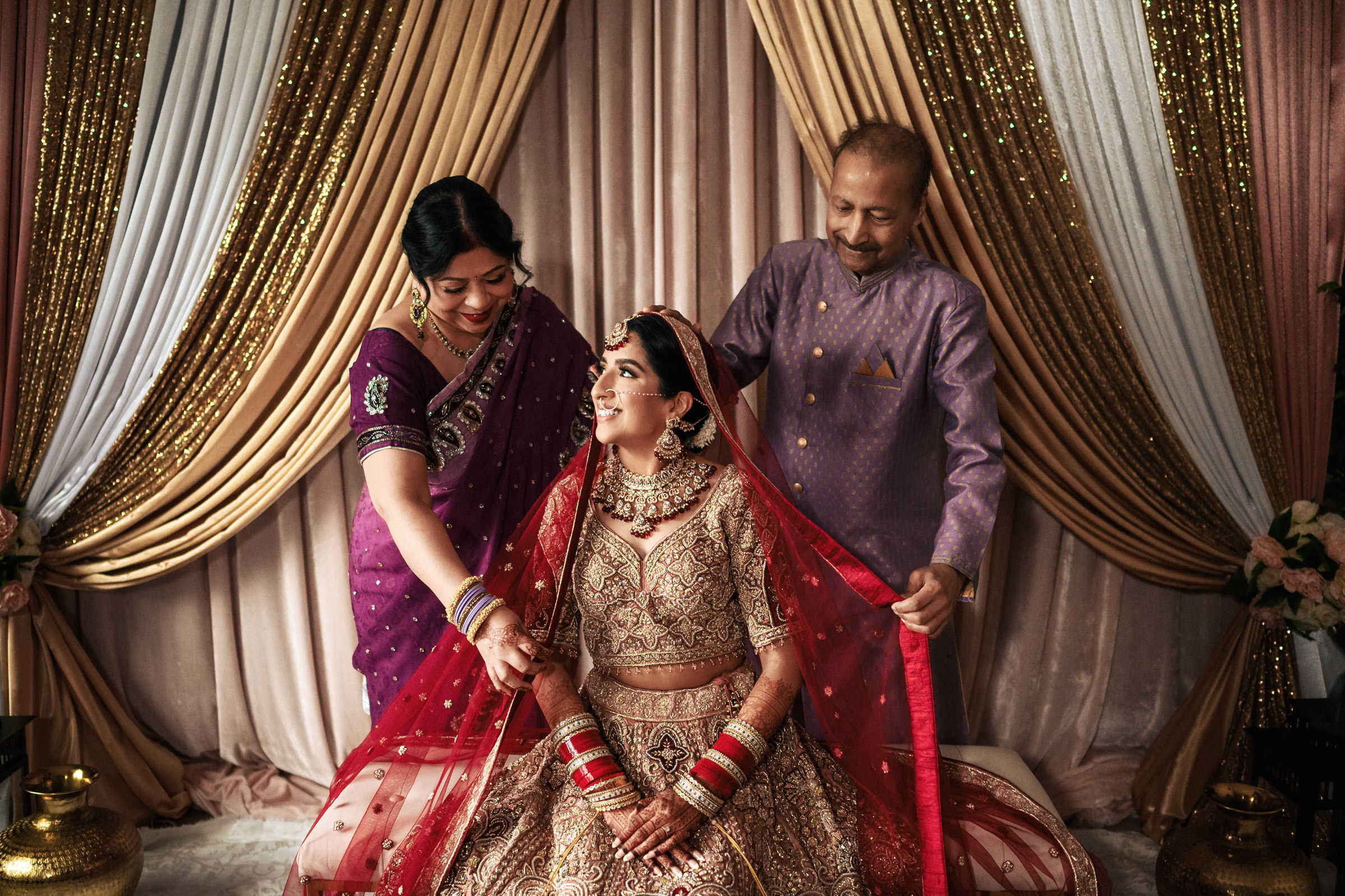 A bride, adorned in a traditional red and gold bridal outfit, sits smiling as her parents stand beside her, adjusting her veil. The background features draped golden and white curtains. The parents look joyful, sharing this intimate moment with their daughter.