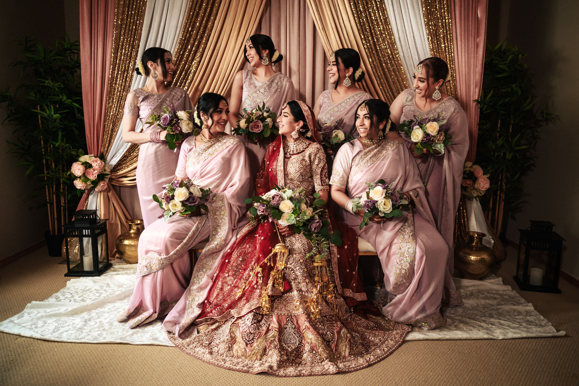 A bride in an intricately embroidered red and gold dress sits surrounded by six bridesmaids in matching lavender sarees. Each bridesmaid holds a bouquet of roses, and they all smile and laugh together. The backdrop features draped fabrics in gold, pink, and cream.