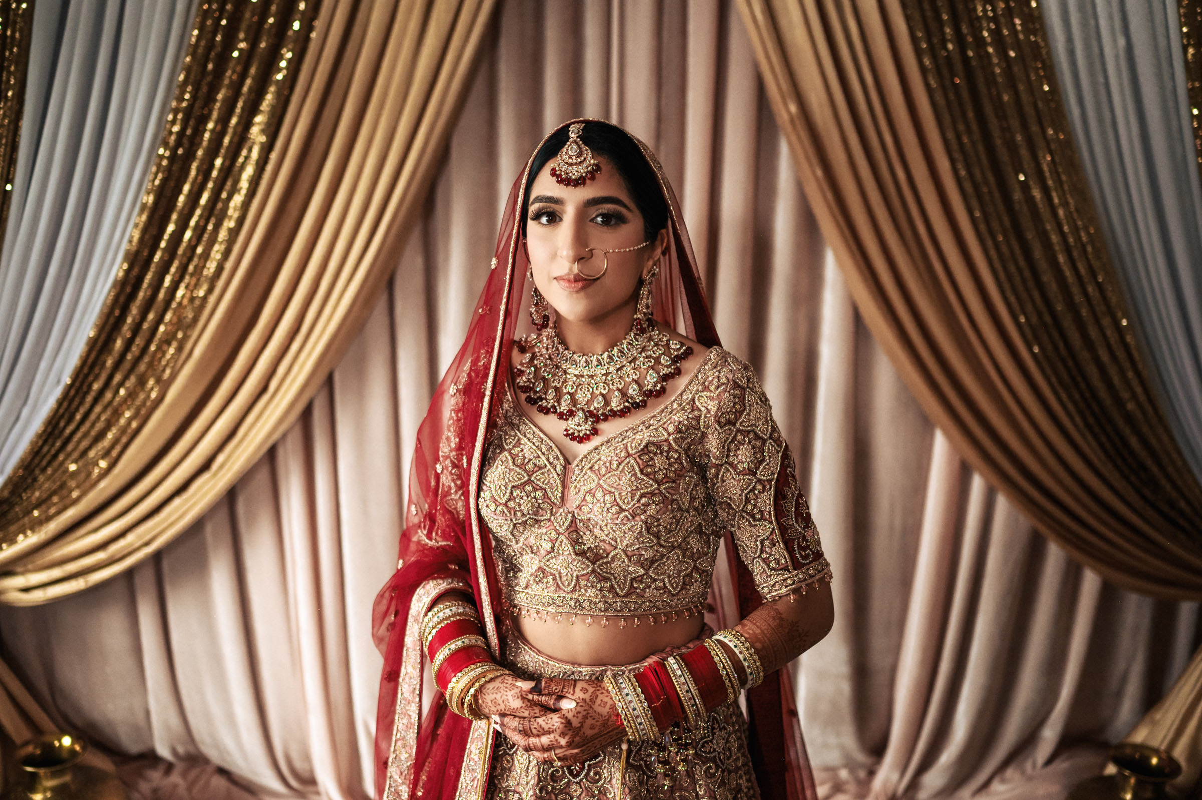 A woman dressed in traditional South Asian bridal attire stands in front of golden drapes. She wears a rich red and gold embroidered outfit, a veil, ornate jewelry, and henna on her hands. She gazes calmly at the camera, displaying elegance and grace.