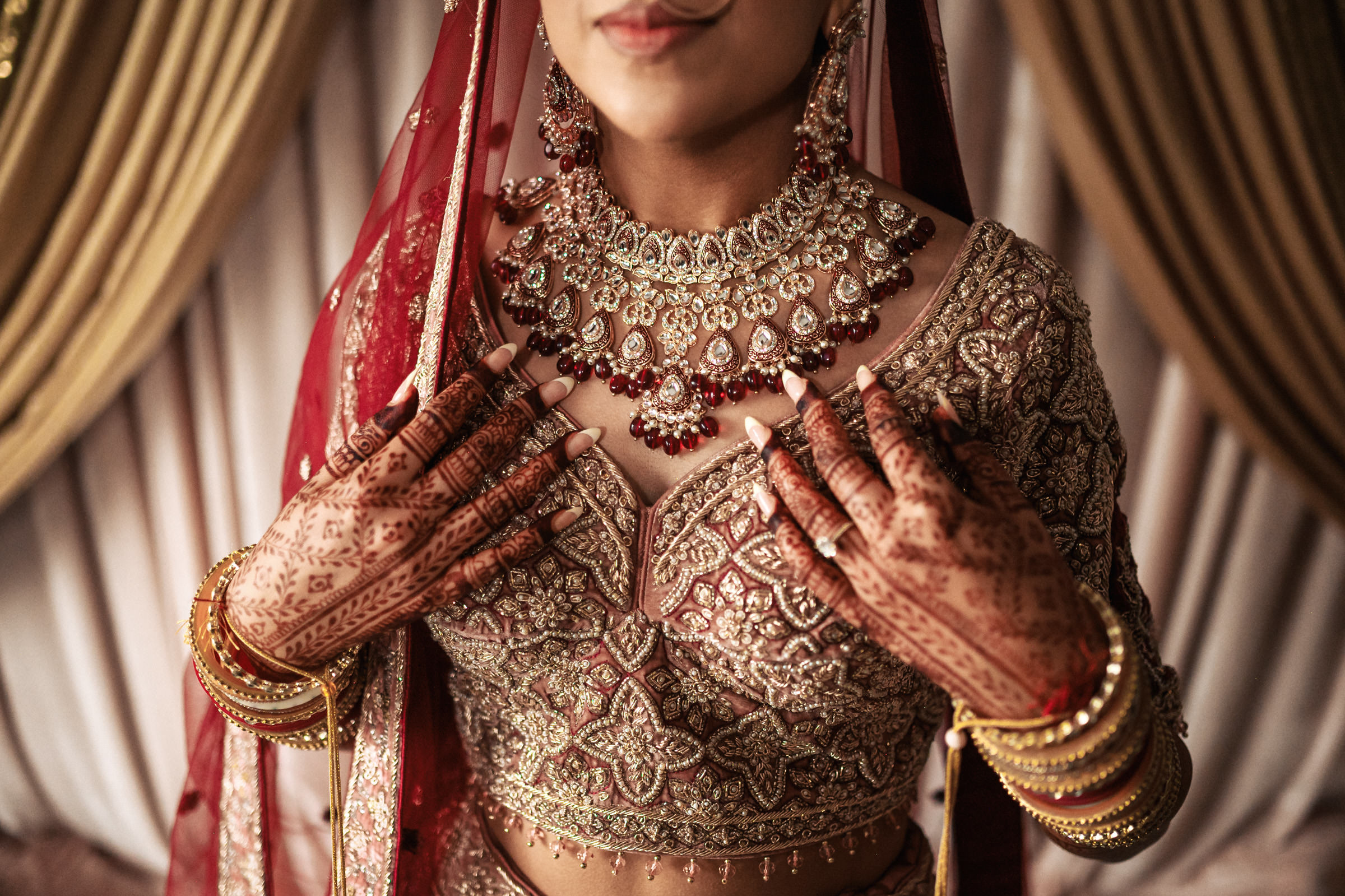 A bride adorned in a richly embellished red bridal outfit, detailed with intricate embroidery, gazes downwards. Her hands, decorated with henna, gently hold her ornate necklace, which matches her elaborate earrings. Gold bangles adorn her wrists. Elegant curtains frame the background.