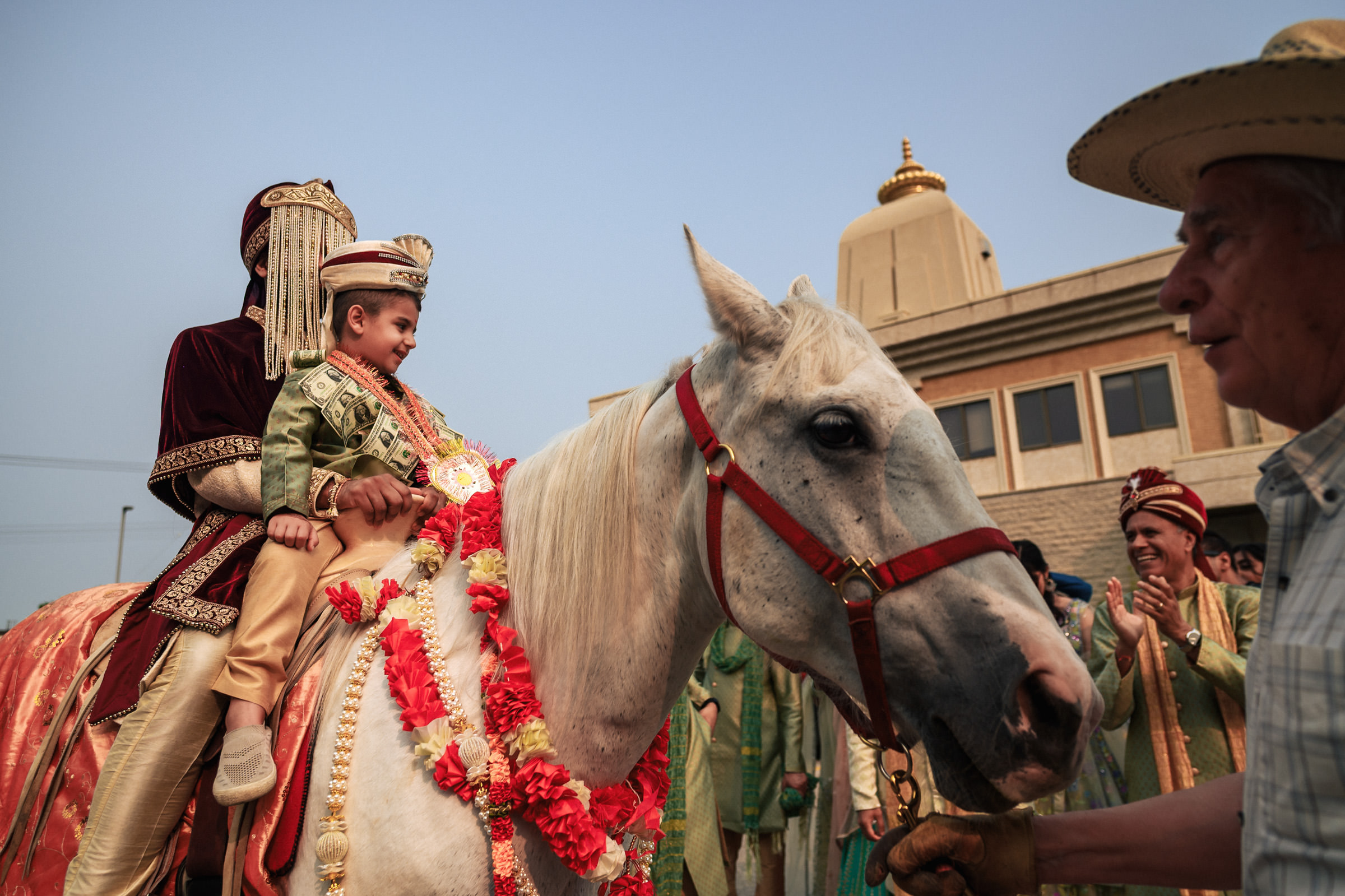 A young boy dressed in traditional ceremonial attire sits atop a decorated white horse, accompanied by an adult, likely during a wedding procession. They are surrounded by people in festive clothing, with a temple-like building in the background.