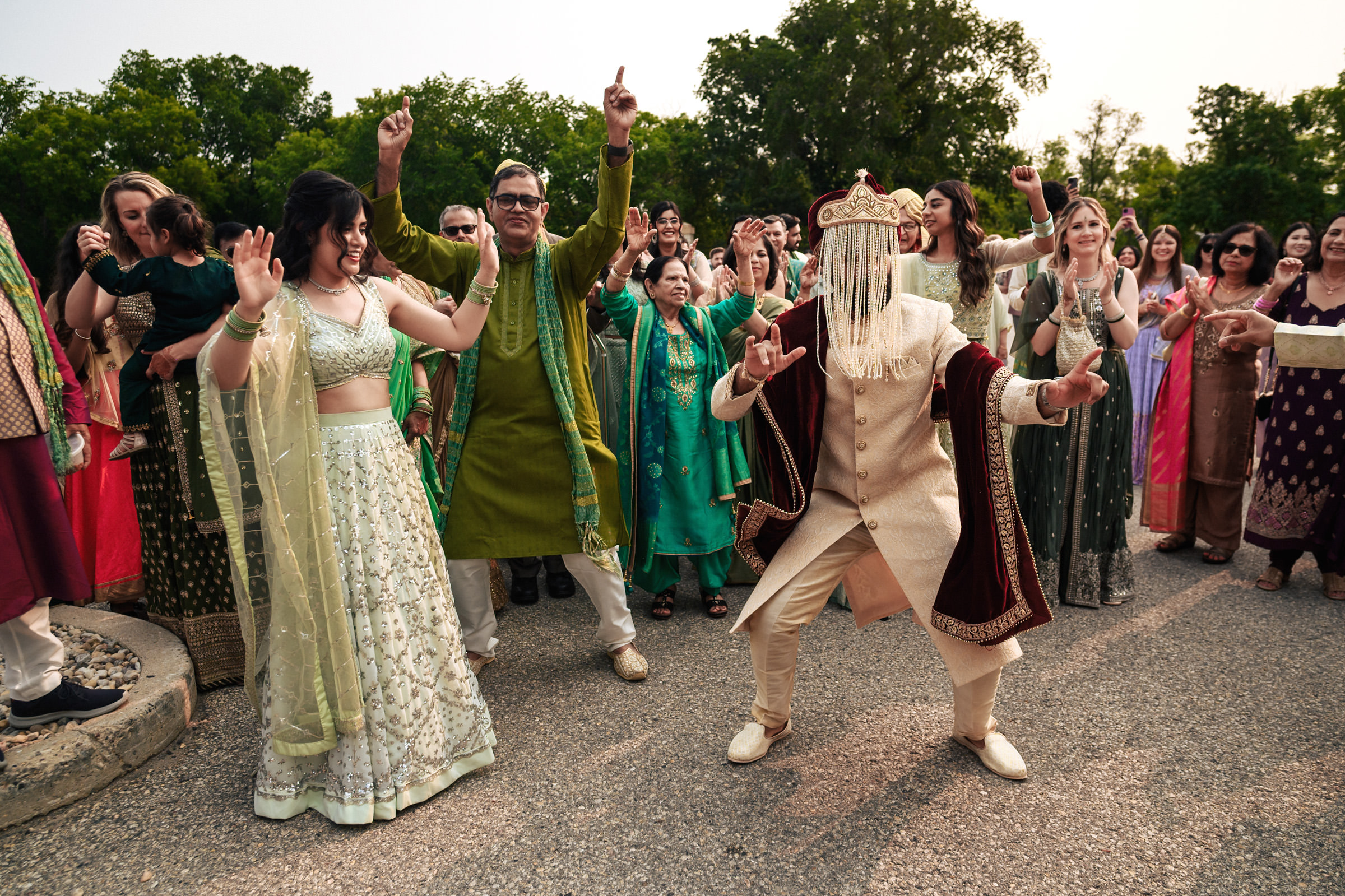 A group of people dressed in traditional South Asian attire celebrate outdoors. A person in an elaborate sherwani and turban, their face covered with a beaded veil, dances energetically in the center. Others surrounding them are dancing and smiling.