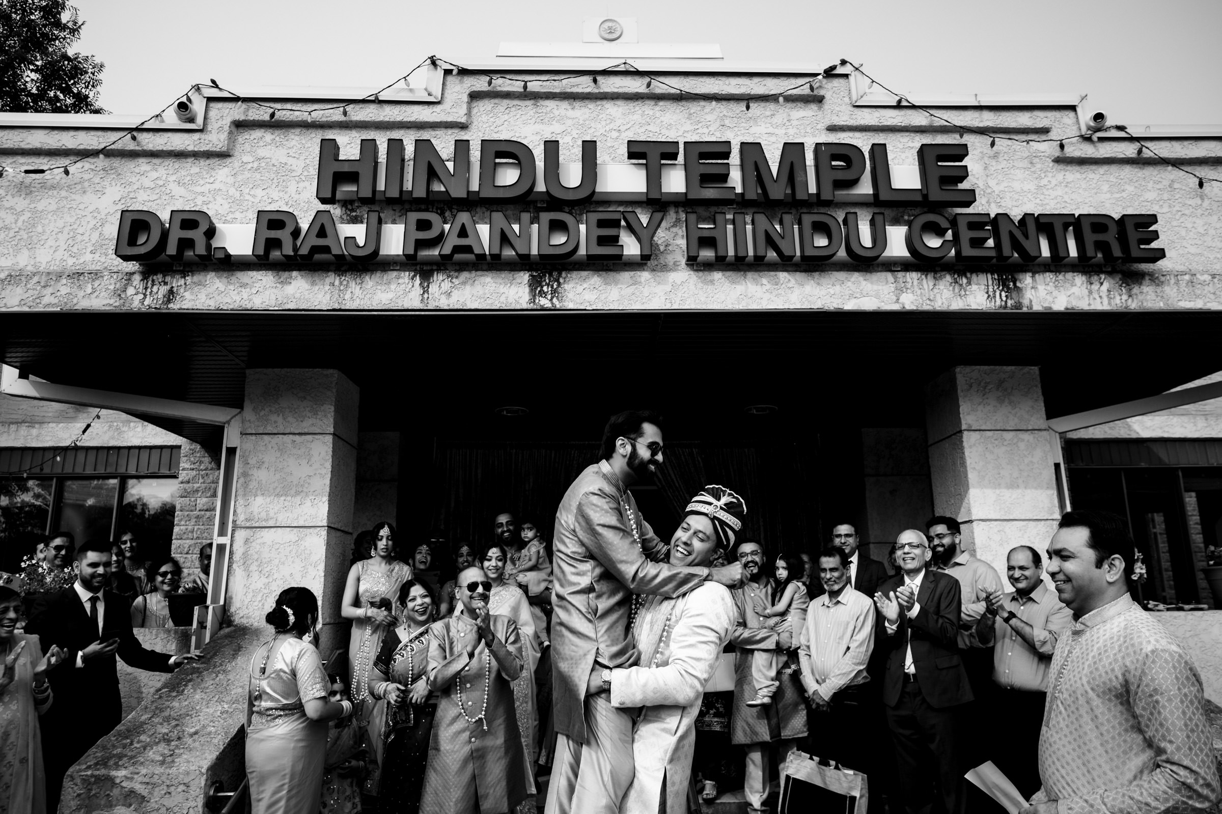 A joyous moment captured in black and white outside a Hindu temple. A groom lifts another man in a celebratory hug, surrounded by smiling and clapping guests dressed in traditional attire. The temple sign reads "Dr. Raj Pandey Hindu Centre.