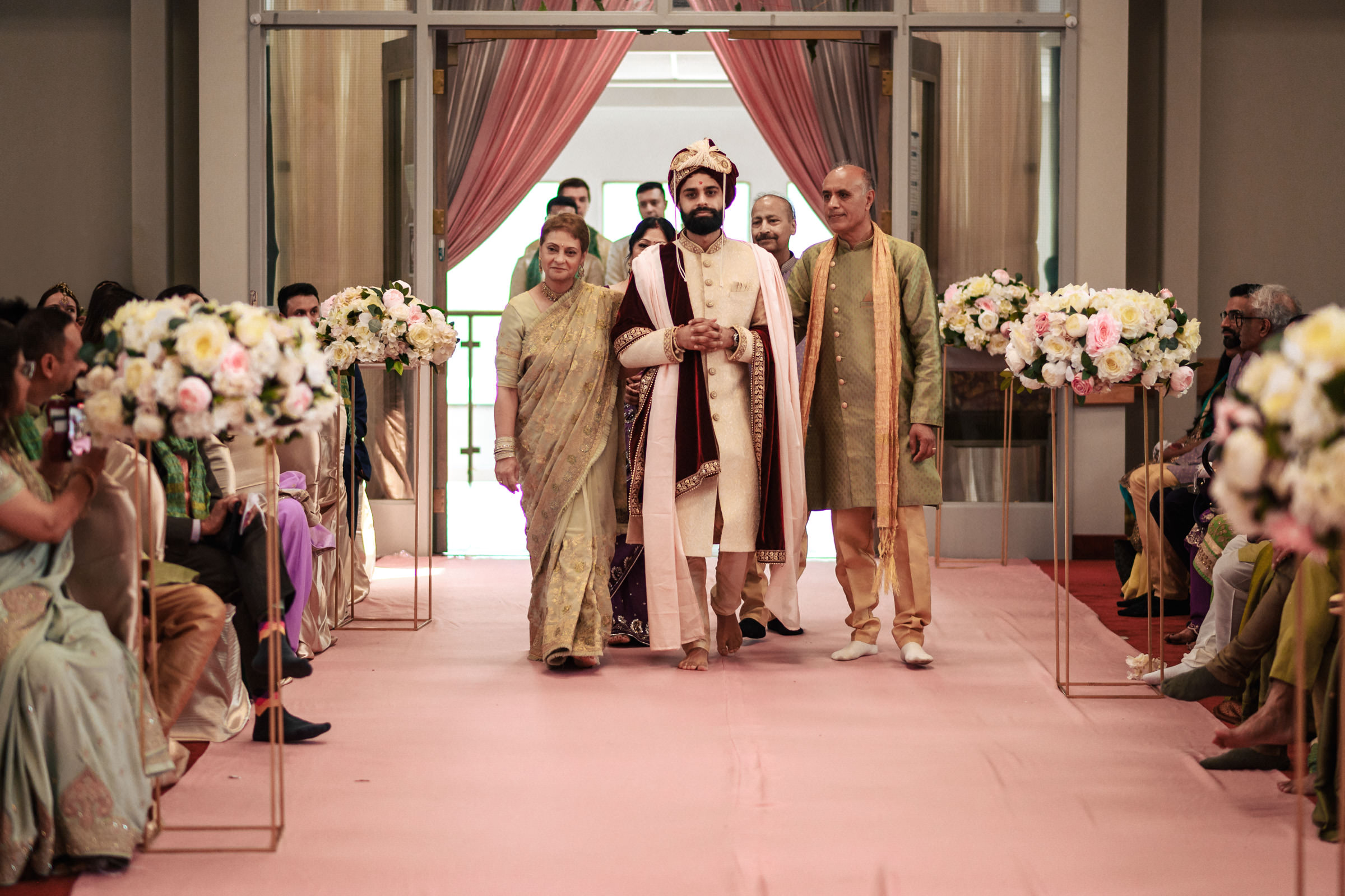 A groom dressed in traditional attire walks down the aisle with two older adults, possibly his parents, beside him. They are in a decorated hall with floral arrangements lining the aisle. Guests are seated on either side, watching the procession.