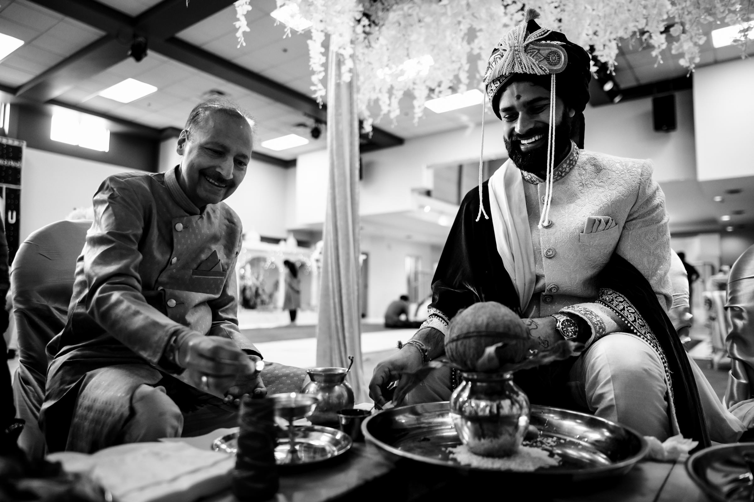 A groom, dressed in traditional attire and a turban, sits smiling beside an older man during a wedding ceremony. Both are engaged in rituals, surrounded by ceremonial items on trays. The setting is indoors, with floral decorations hanging from the ceiling.