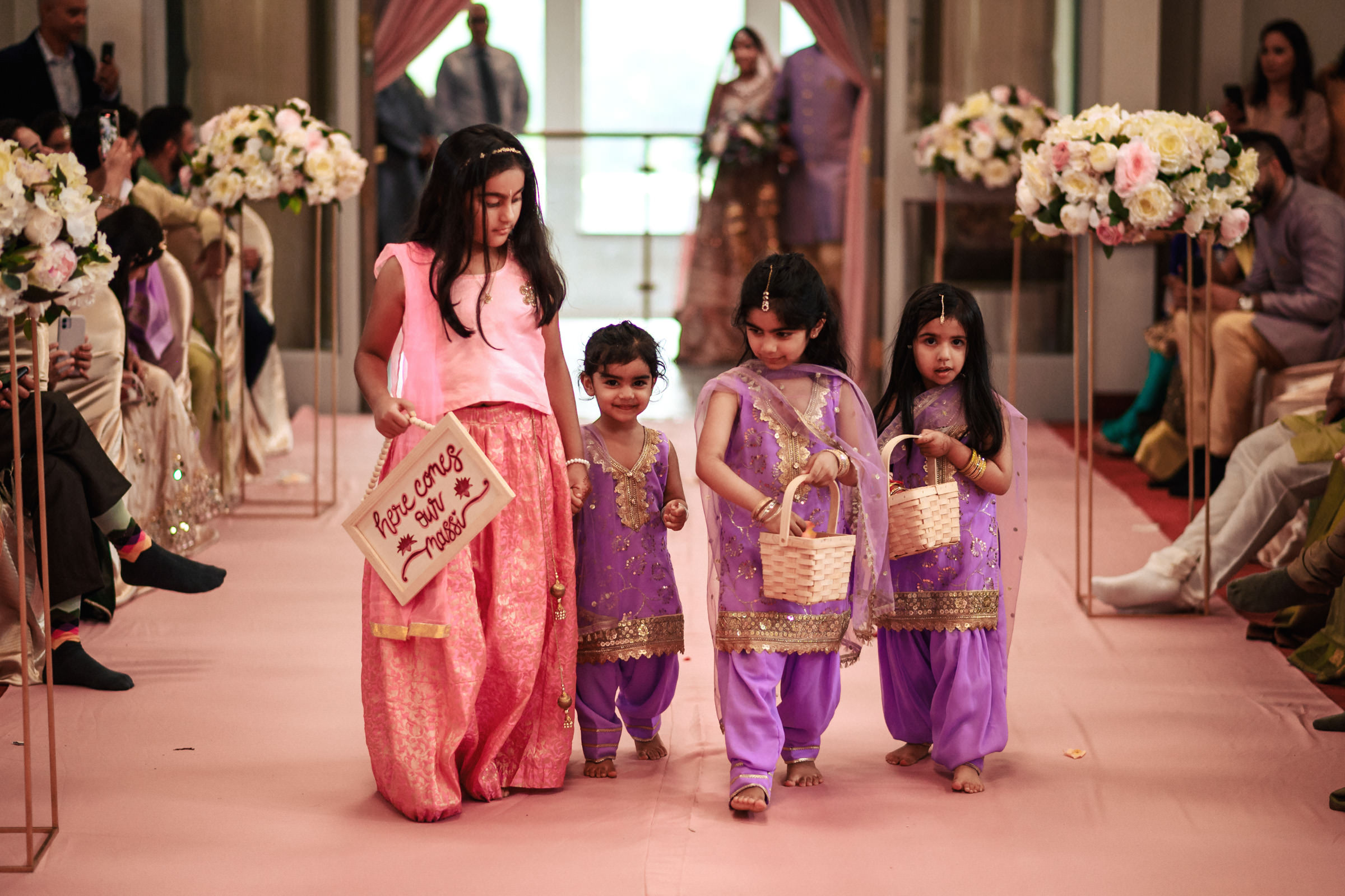 Four children are walking down a decorated aisle at a wedding. The leading girl holds a sign that reads, "Here comes the bride!" The other three, dressed in purple outfits, carry small baskets. Guests on either side watch and smile. Floral arrangements adorn the aisle.