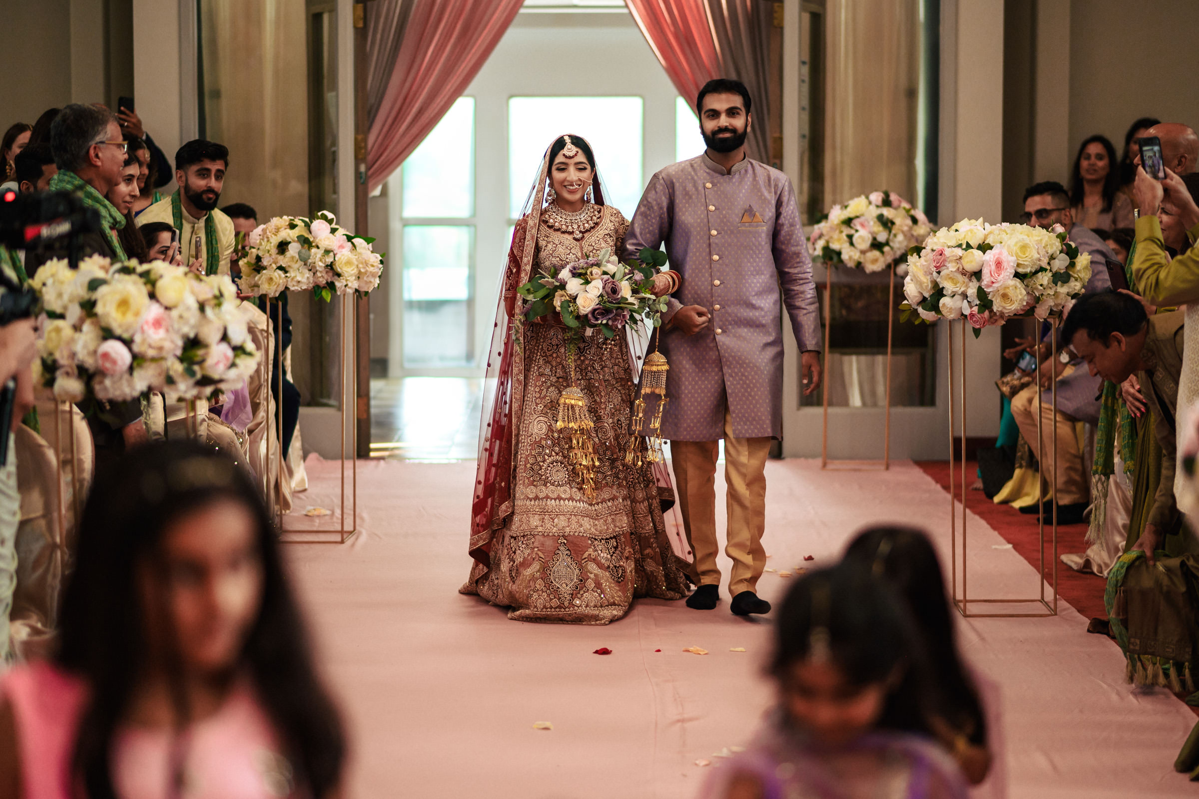 A bride and groom in traditional wedding attire walk down an aisle adorned with flowers, surrounded by guests in colorful outfits. The bride wears an intricate red and gold dress, while the groom dons a lavender sherwani. The backdrop includes draped curtains and floral arrangements.