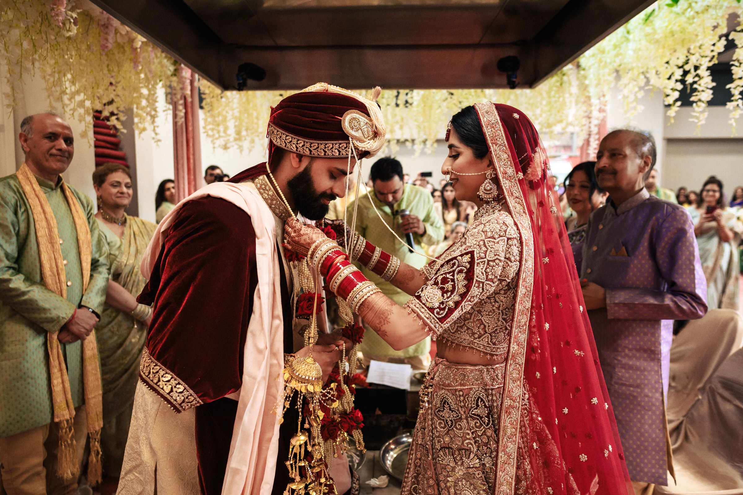 A bride and groom, dressed in traditional Indian wedding attire, participate in a ceremony. The bride adjusts the groom's necklace. They are surrounded by family and friends, who watch with smiles. Floral decorations and greenery are draped around the ceremonial area.
