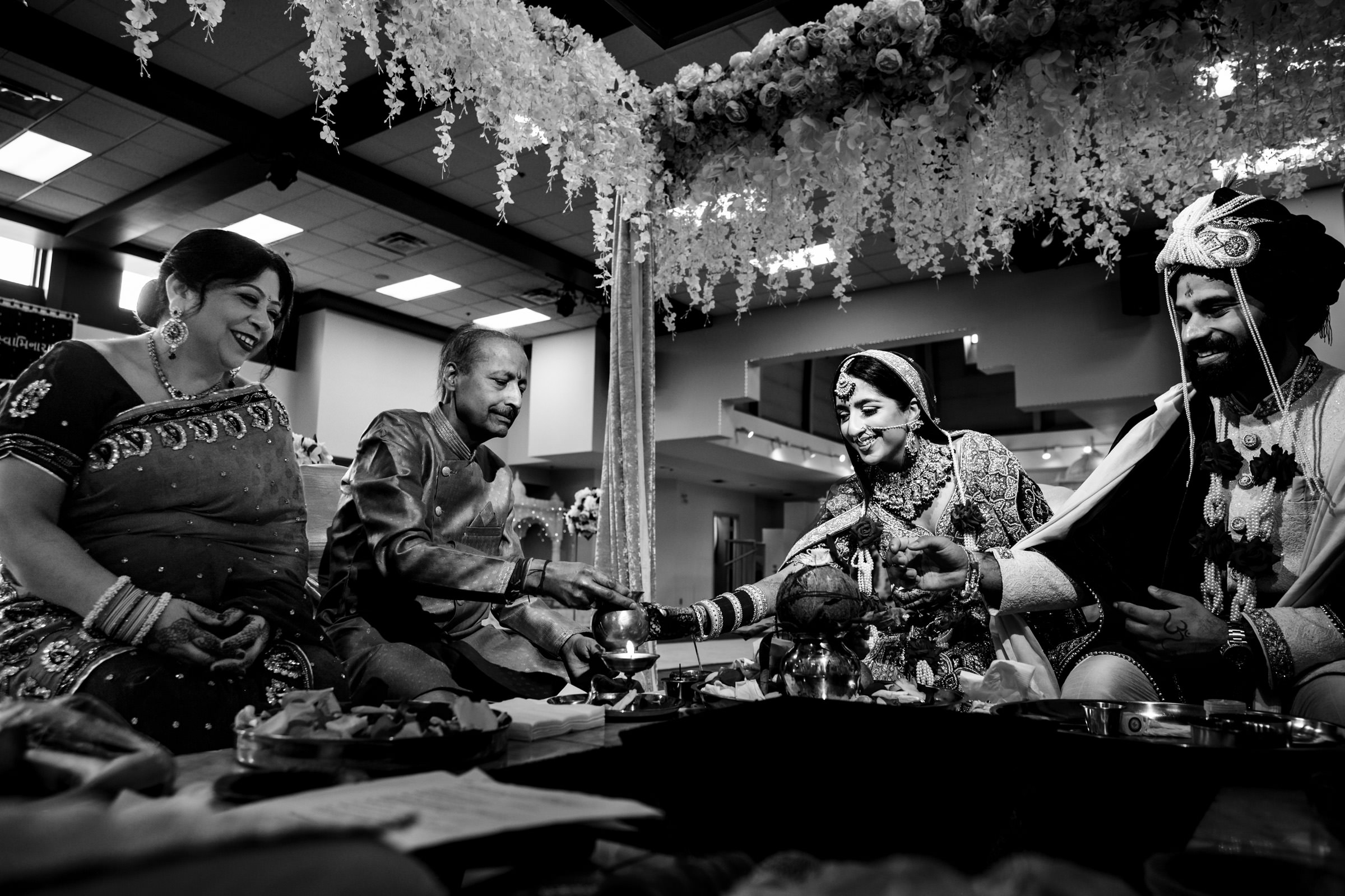 A black-and-white photo of a couple's wedding ceremony. The bride and groom, dressed in traditional attire, are seated and smiling while performing rituals. Two seated guests, also in traditional clothes, are smiling and watching the ceremony. Floral decorations hang overhead.