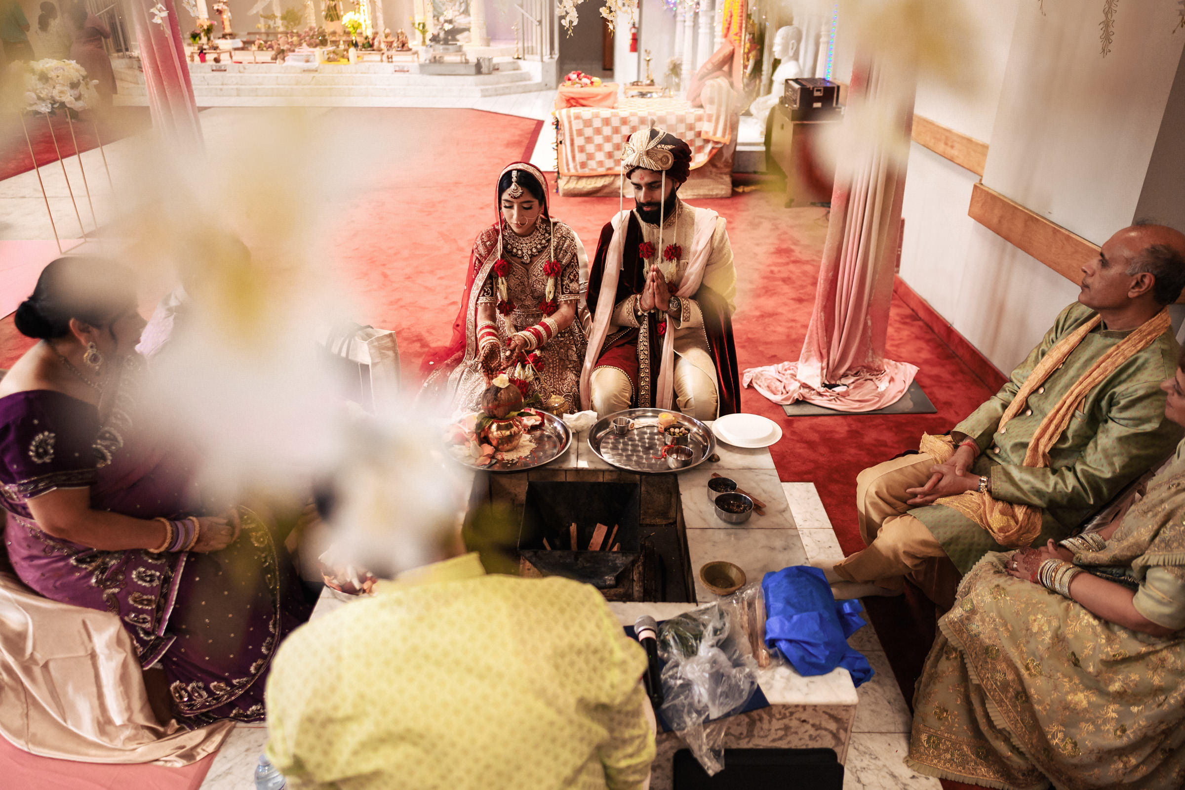 A bride and groom sit side by side in traditional wedding attire during a ceremony, surrounded by family members. They are performing a ritual involving fire. The setting is ornate, with red carpets and decorative elements in the background.