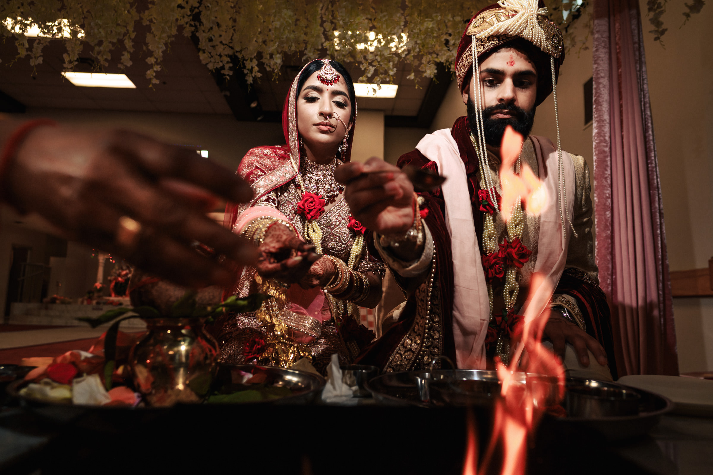 A bride and groom, dressed in traditional Indian wedding attire, perform a ritual at a Hindu wedding ceremony. They hold offerings over a sacred fire, with flowers and ceremonial items surrounding them. The atmosphere is solemn and sacred.
