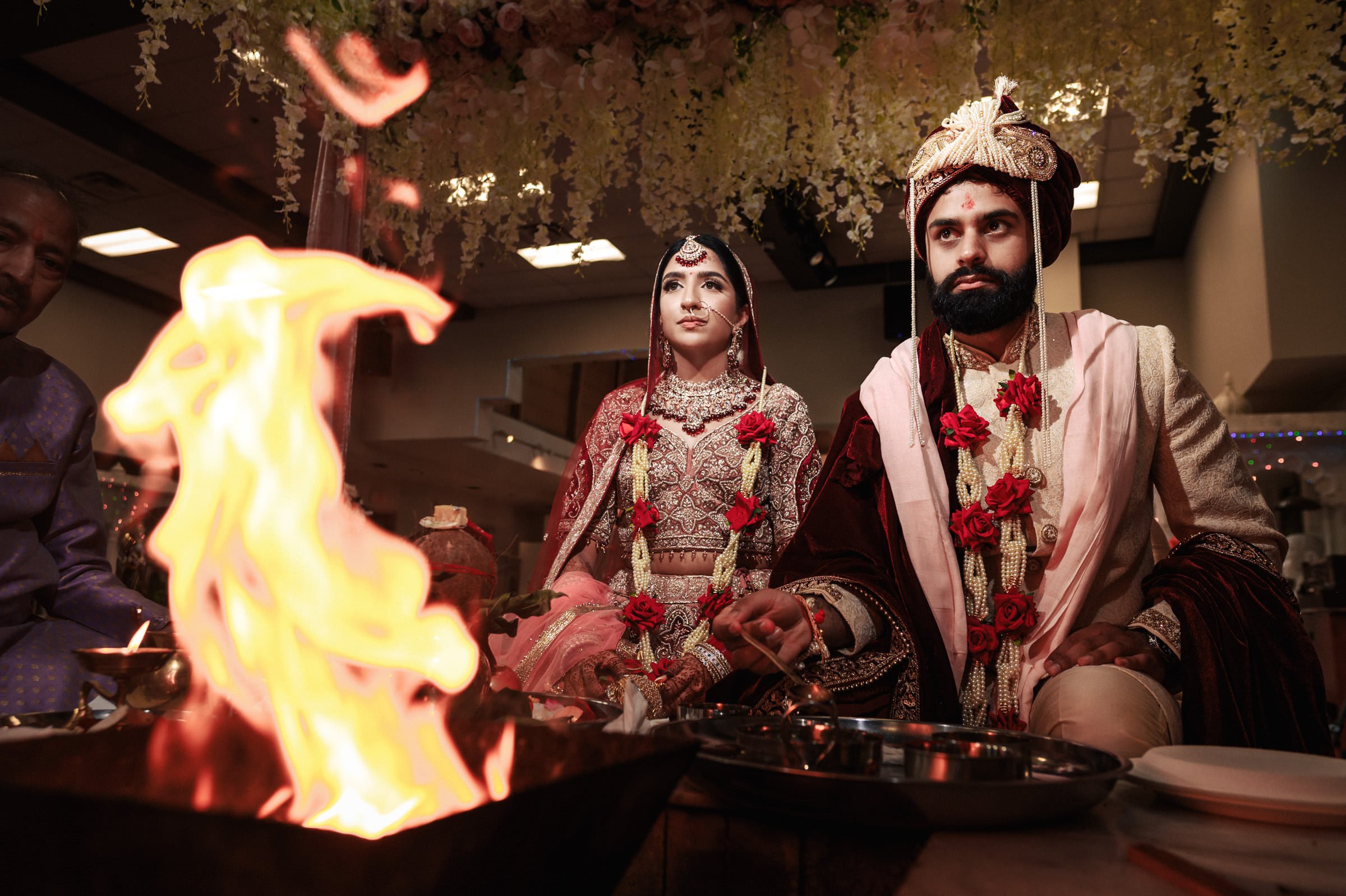 A bride and groom, dressed in traditional Indian wedding attire, sit side by side during a wedding ceremony. The groom wears a turban and the bride wears ornate jewelry. They are sitting in front of a fire ritual, with flowers hanging above them.