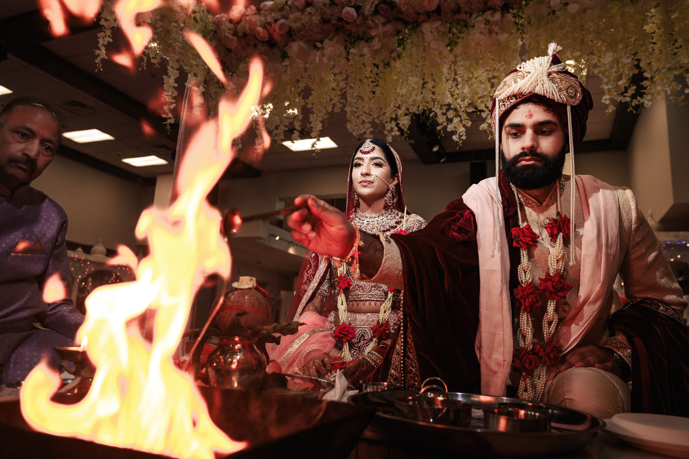 A couple in traditional attire participates in a Hindu wedding ritual. The groom, wearing a turban and garlands, makes offerings into a ceremonial fire. The bride, adorned with jewelry and a veil, looks on. Floral decorations hang above them.