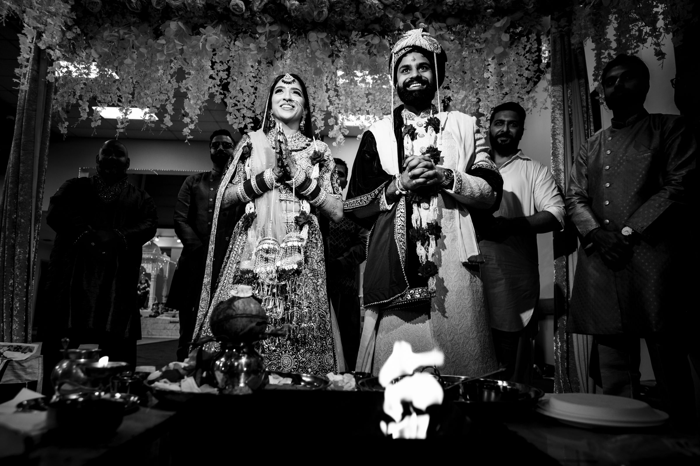 A black and white image depicting a joyful couple in traditional attire standing under a floral canopy during a wedding ceremony. They are surrounded by smiling guests, with ceremonial items and a small fire visible in the foreground.