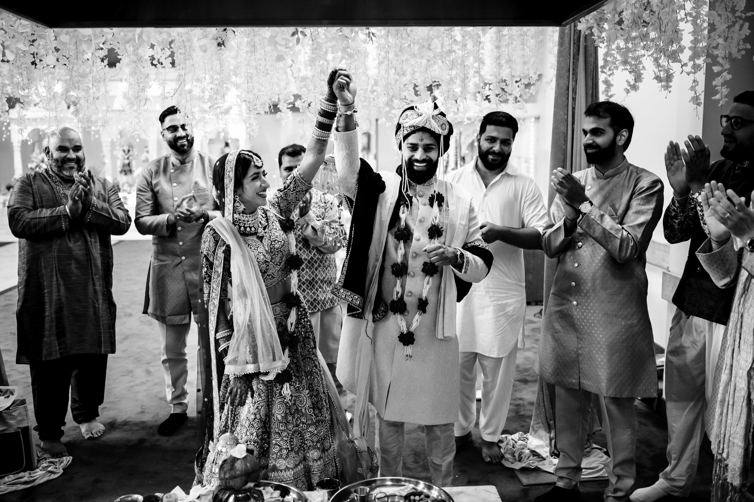 A joyful couple celebrates their wedding, raising their hands together. The bride is in an ornate traditional dress and the groom in traditional attire with a turban. They are surrounded by smiling, clapping friends and family, all dressed in elegant traditional clothing.
