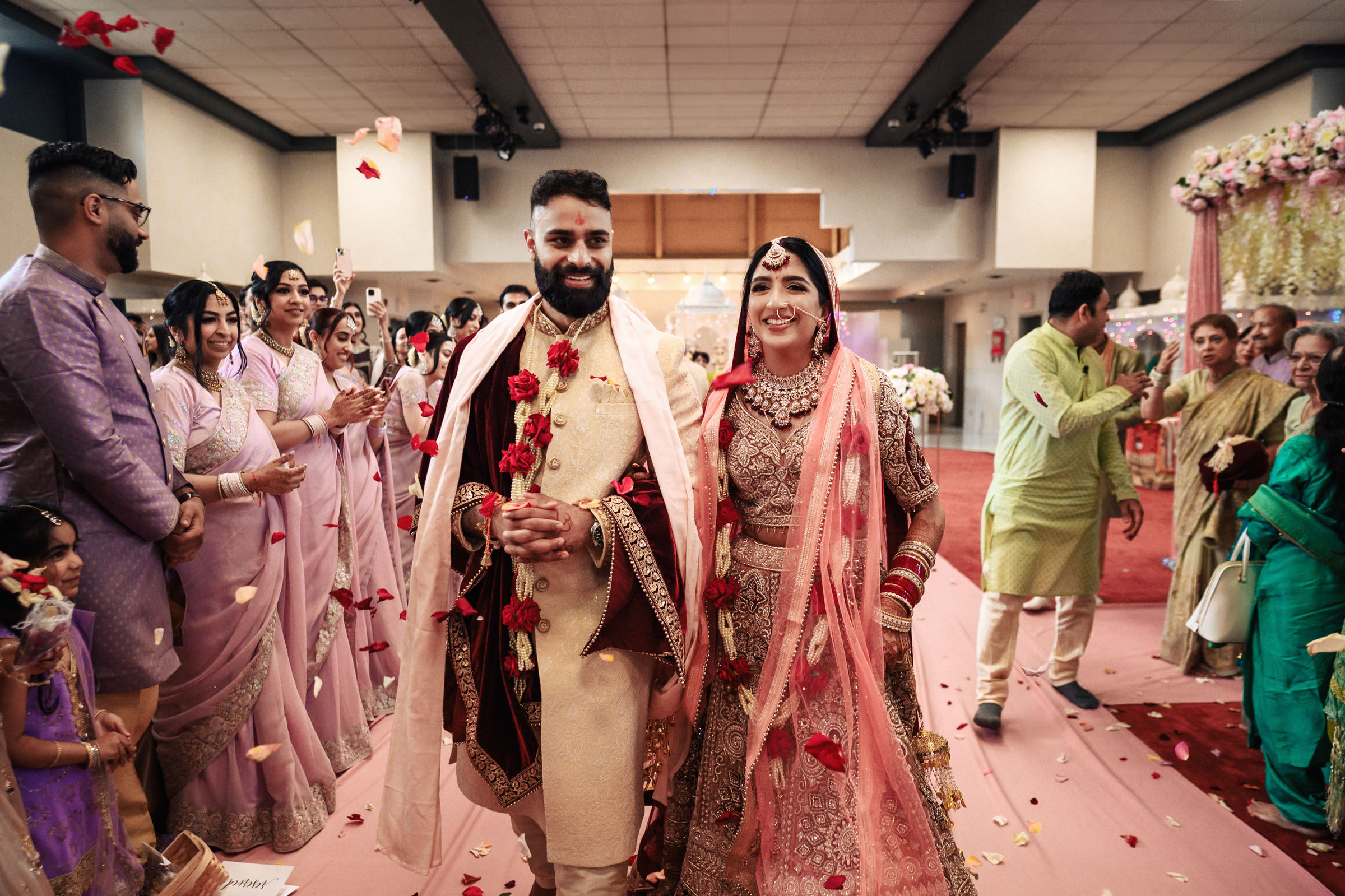 A couple in traditional attire joyfully walks down an aisle as flower petals fall around them, surrounded by smiling guests who are dressed in colorful clothes. The background features elegant decorations, hinting at a wedding ceremony in a beautifully adorned hall.