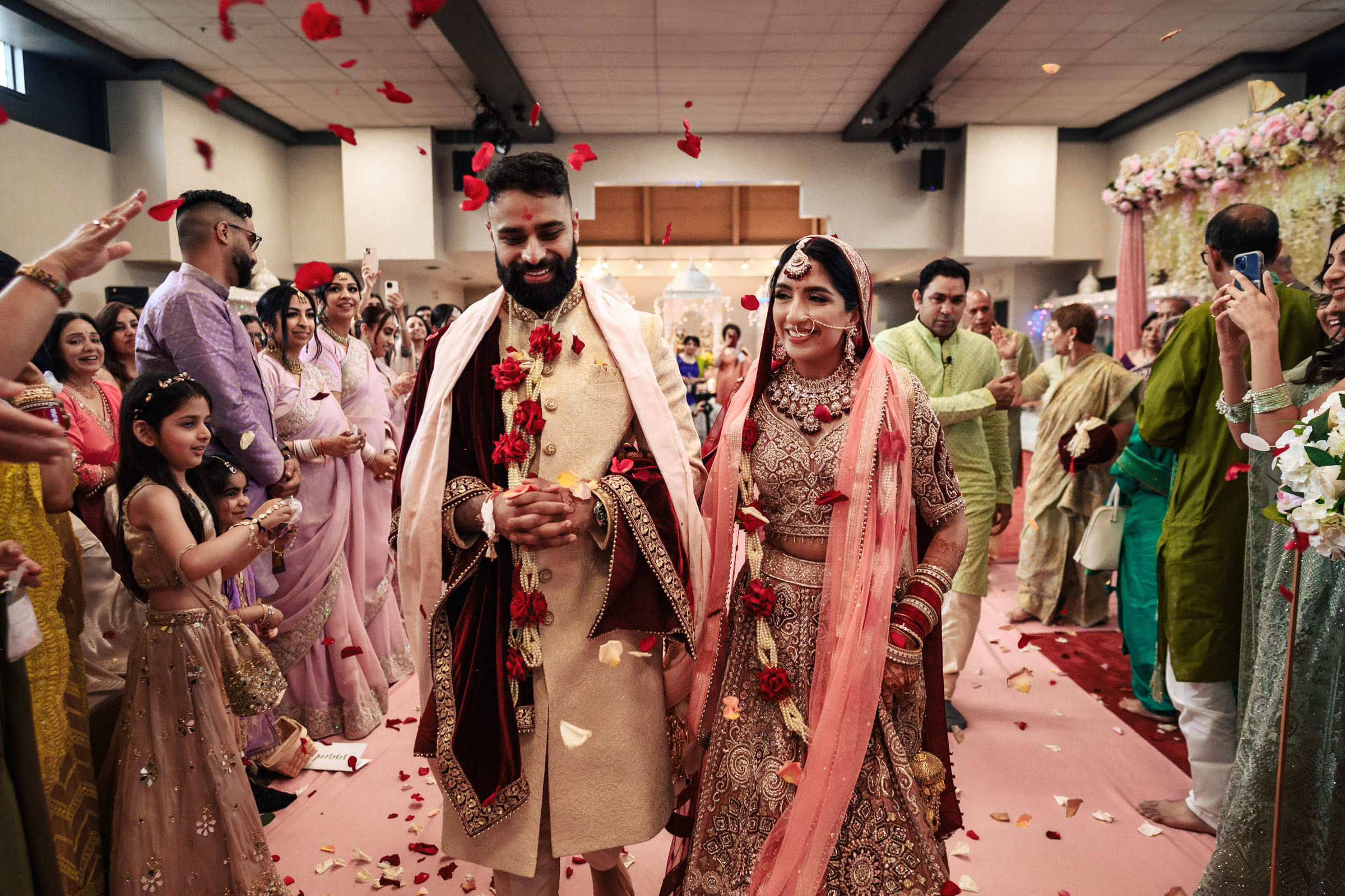 A couple in traditional attire walks down the aisle after their wedding ceremony, surrounded by joyful guests who are showering them with rose petals. The bride wears an ornate lehenga and veil, while the groom is in a sherwani. The atmosphere is festive and vibrant.