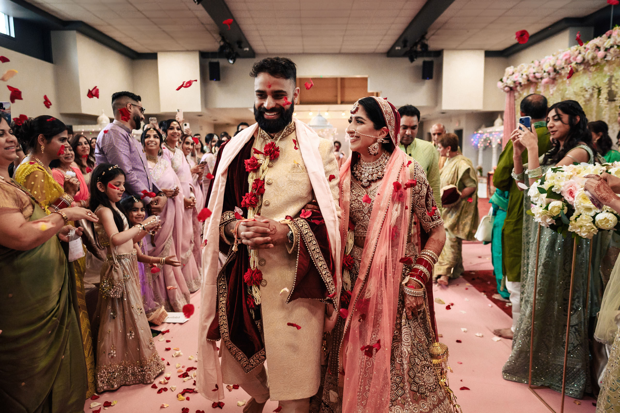 A joyous wedding scene featuring a smiling bride and groom in traditional Indian attire walking down an aisle amidst friends and family. The bride wears a pink lehenga and the groom dons a beige sherwani. Rose petals are thrown in celebration as onlookers cheer.