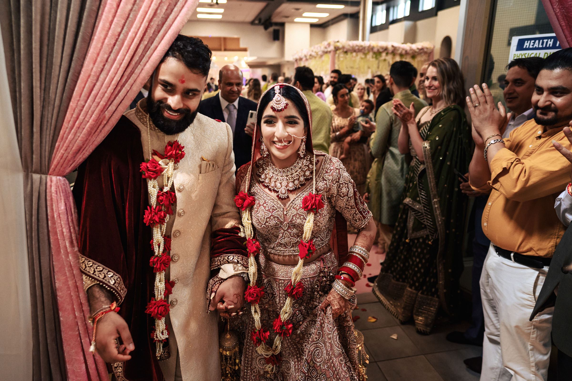A couple dressed in traditional Indian wedding attire holds hands and smiles while walking down a decorated hallway. The bride wears an ornate dress and jewelry, while the groom is in a sherwani. Guests in the background clap and celebrate.