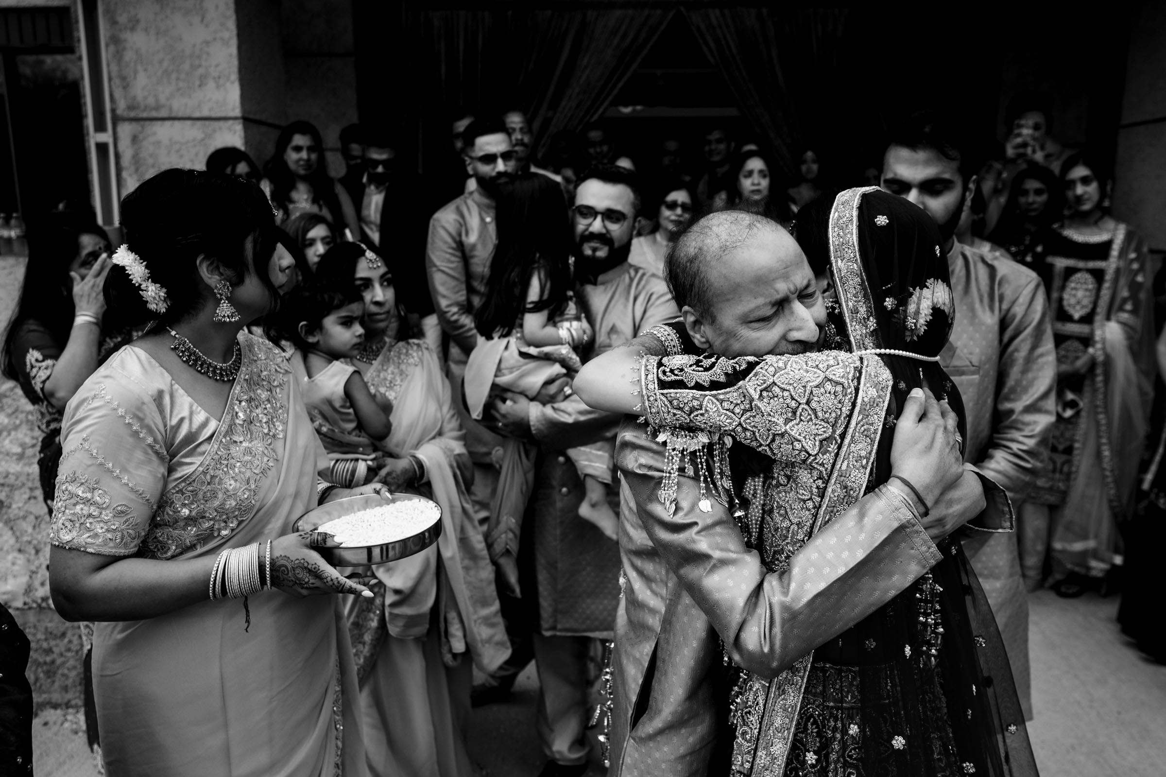 A bride, dressed in traditional attire, embraces an older man tightly. They are surrounded by emotional onlookers, including women in elegant sarees holding plates of rice, seemingly part of a wedding ritual. The scene is filled with joy and sentiment.
