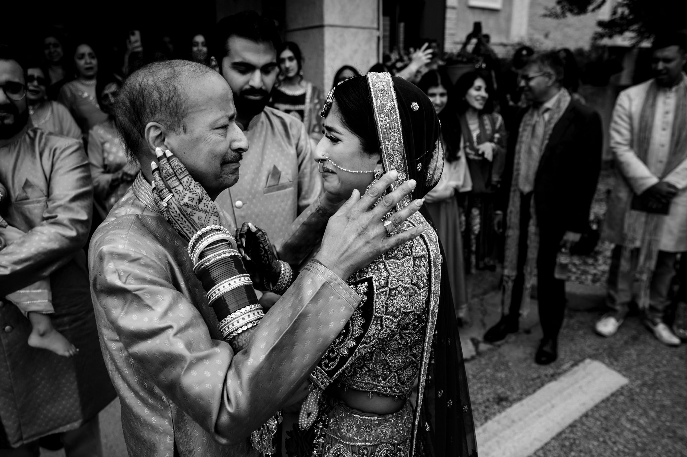 A black-and-white photo of a joyous moment during a wedding. A bride in traditional attire, including a headpiece and bangles, warmly embraces an elderly man, both smiling. Onlookers in traditional clothing share in the celebration in the background.