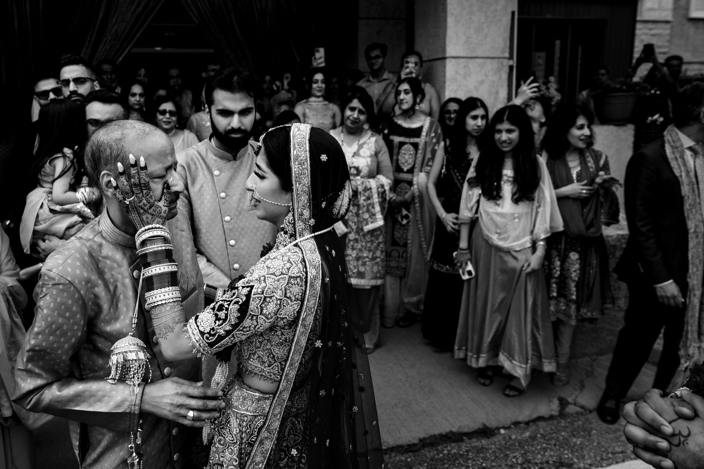 A bride in traditional attire with intricate jewelry and a veil gently touches the face of an older man during what appears to be a wedding ceremony. They are surrounded by an emotional crowd of family and friends, who are watching and taking photos.
