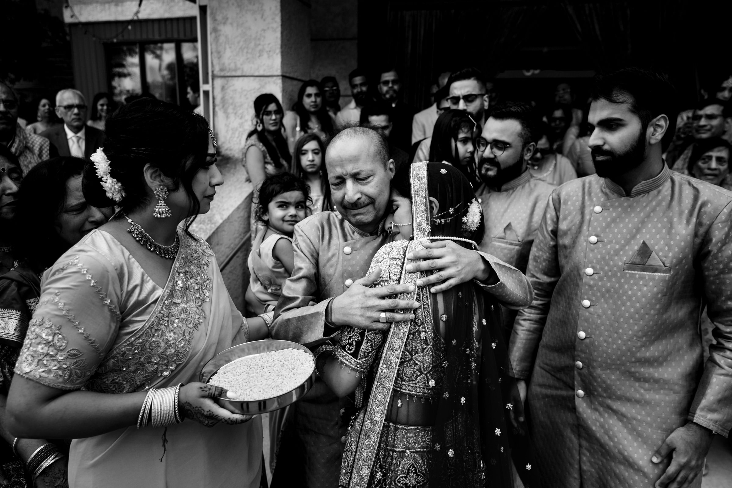A black and white photo capturing an emotional moment at a traditional wedding. The bride, dressed in ornate attire, is embraced by a tearful older man, possibly her father. A woman beside them holds a bowl filled with rice. Onlookers, dressed in traditional clothing, surround them.
