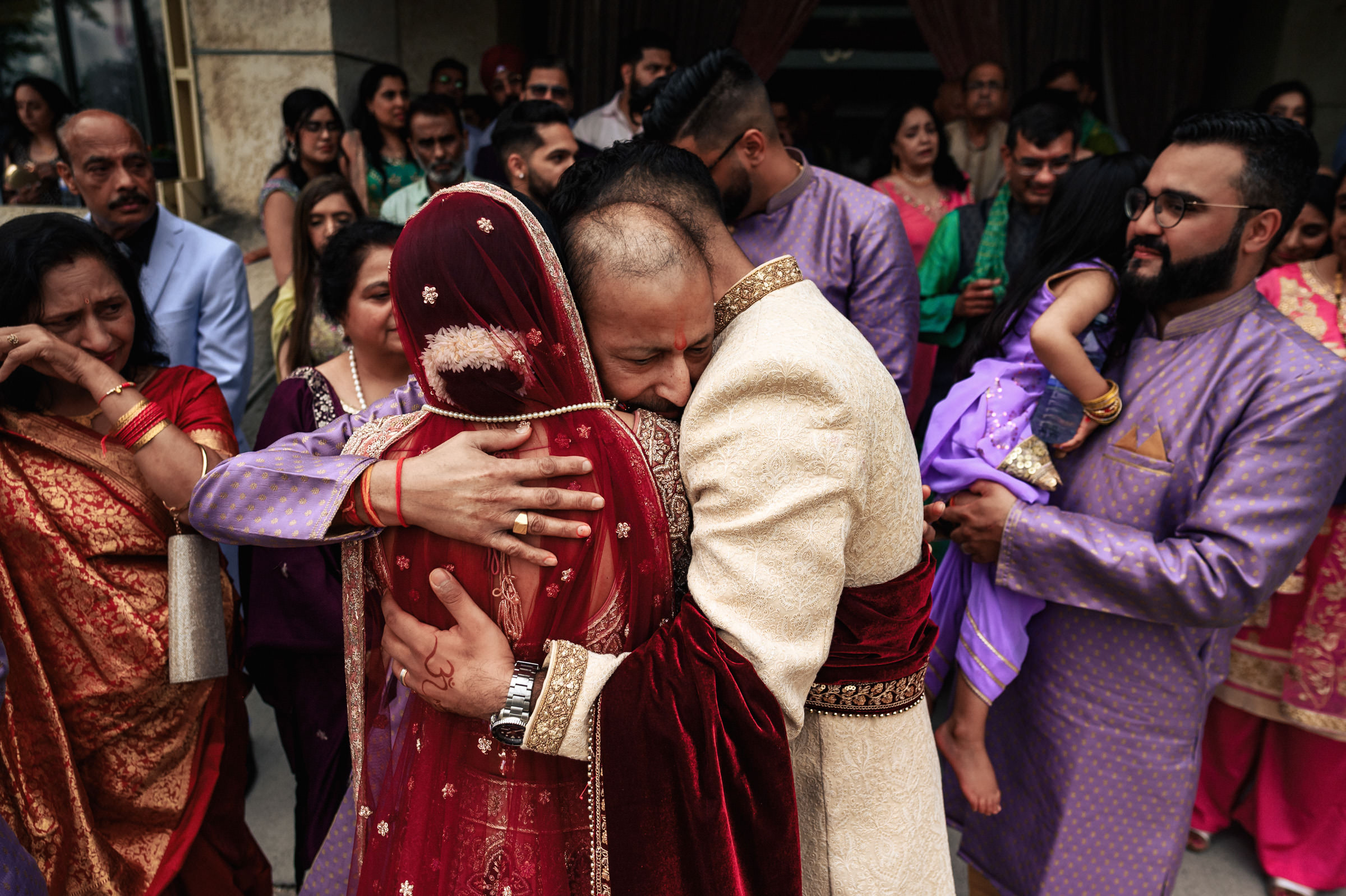 A bride and groom, both dressed in traditional Indian wedding attire, share an emotional hug amid a crowd of family and friends. The groom wears a beige sherwani, and the bride has on a red saree with a veil. The background is filled with people in colorful clothing.