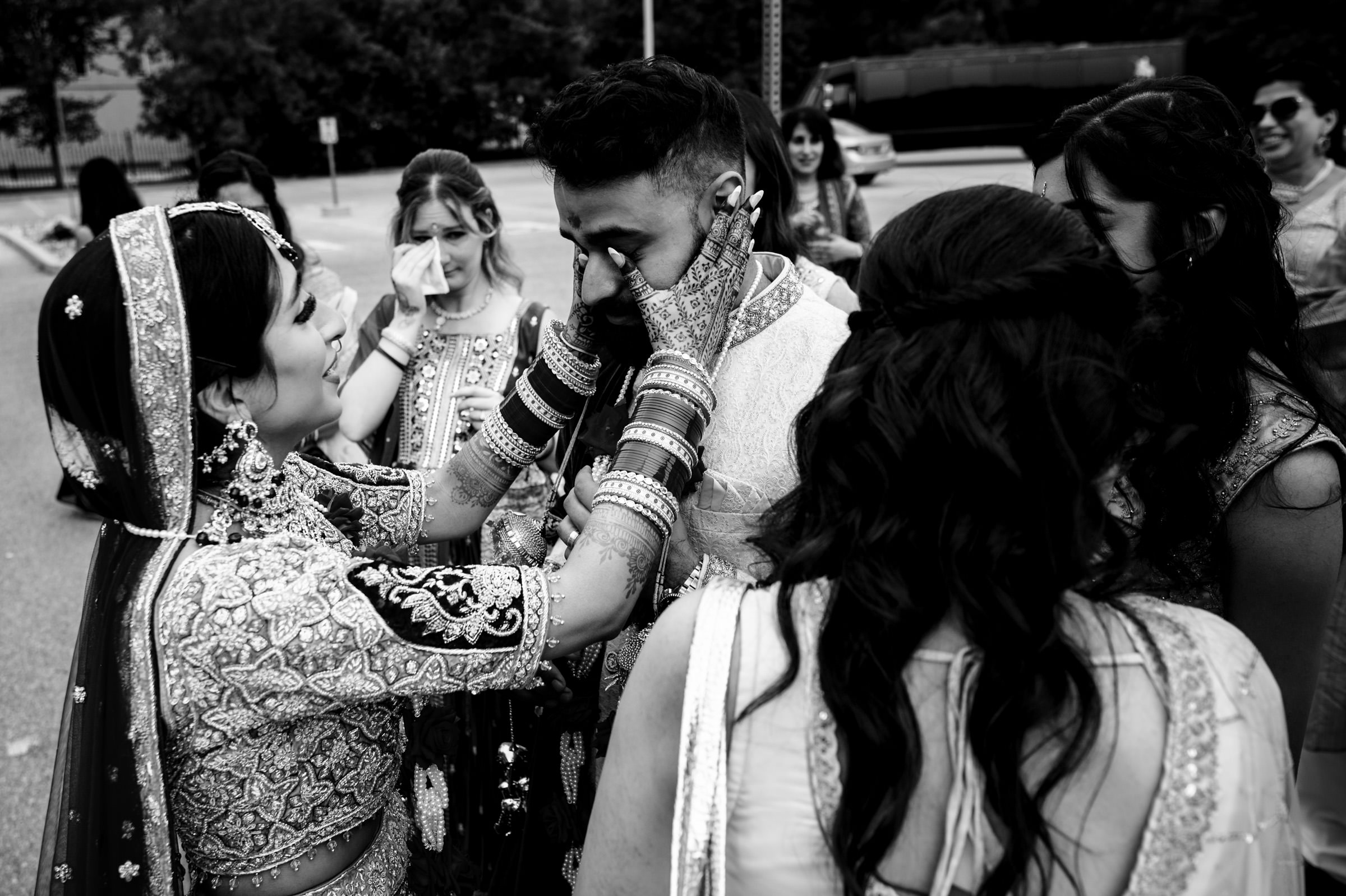A bride, dressed in traditional attire with intricate designs and jewelry, places her hands on the face of a groom who looks emotional. Several bridesmaids in similar attire surround them, some also appearing emotional. The scene is heartfelt and intimate.