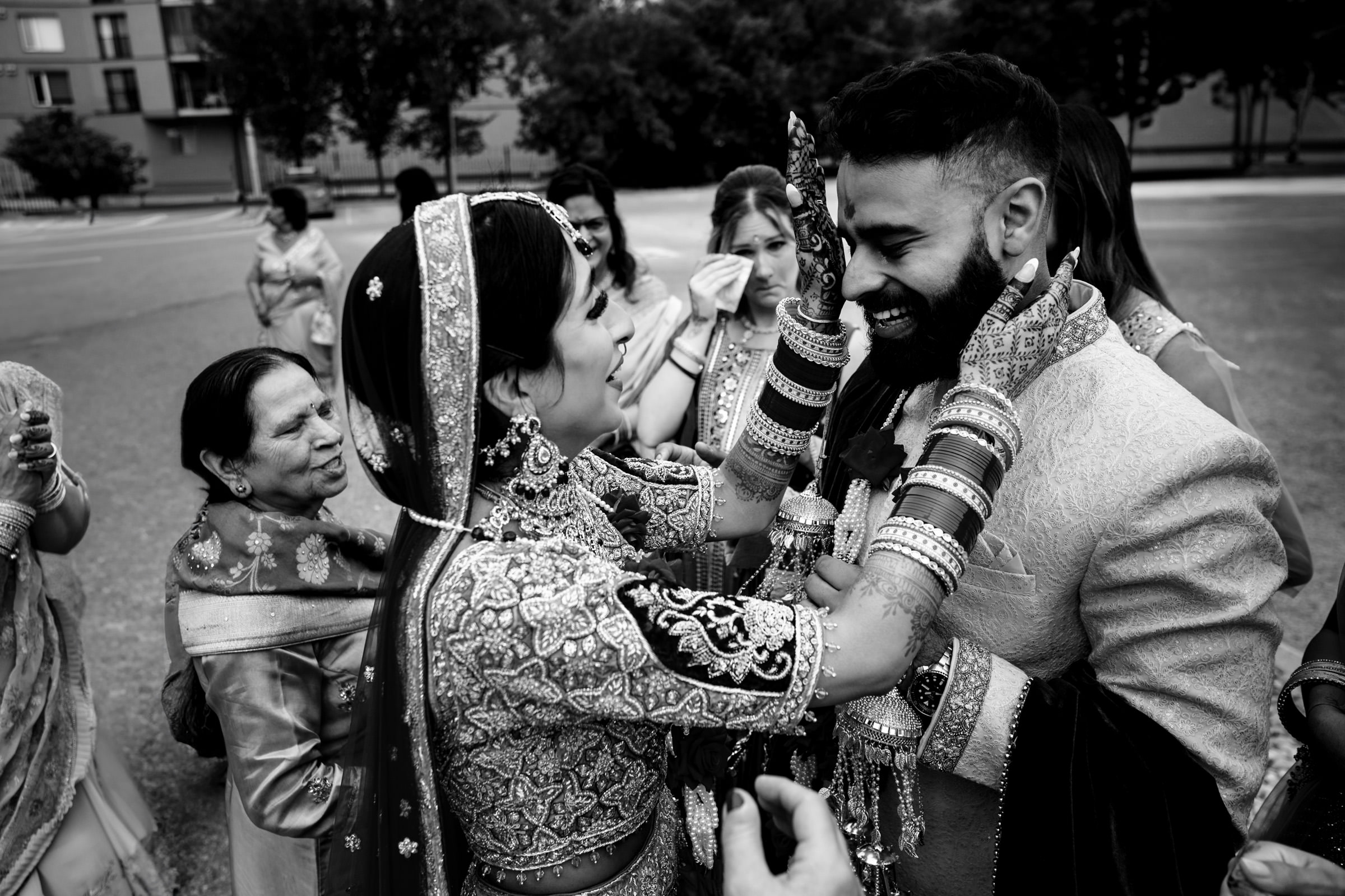 A bride and groom in traditional attire share a joyful moment outdoors, surrounded by family and friends. The bride, smiling, gently holds the groom's face with both hands as the groom grins broadly. The scene exudes happiness and celebration.