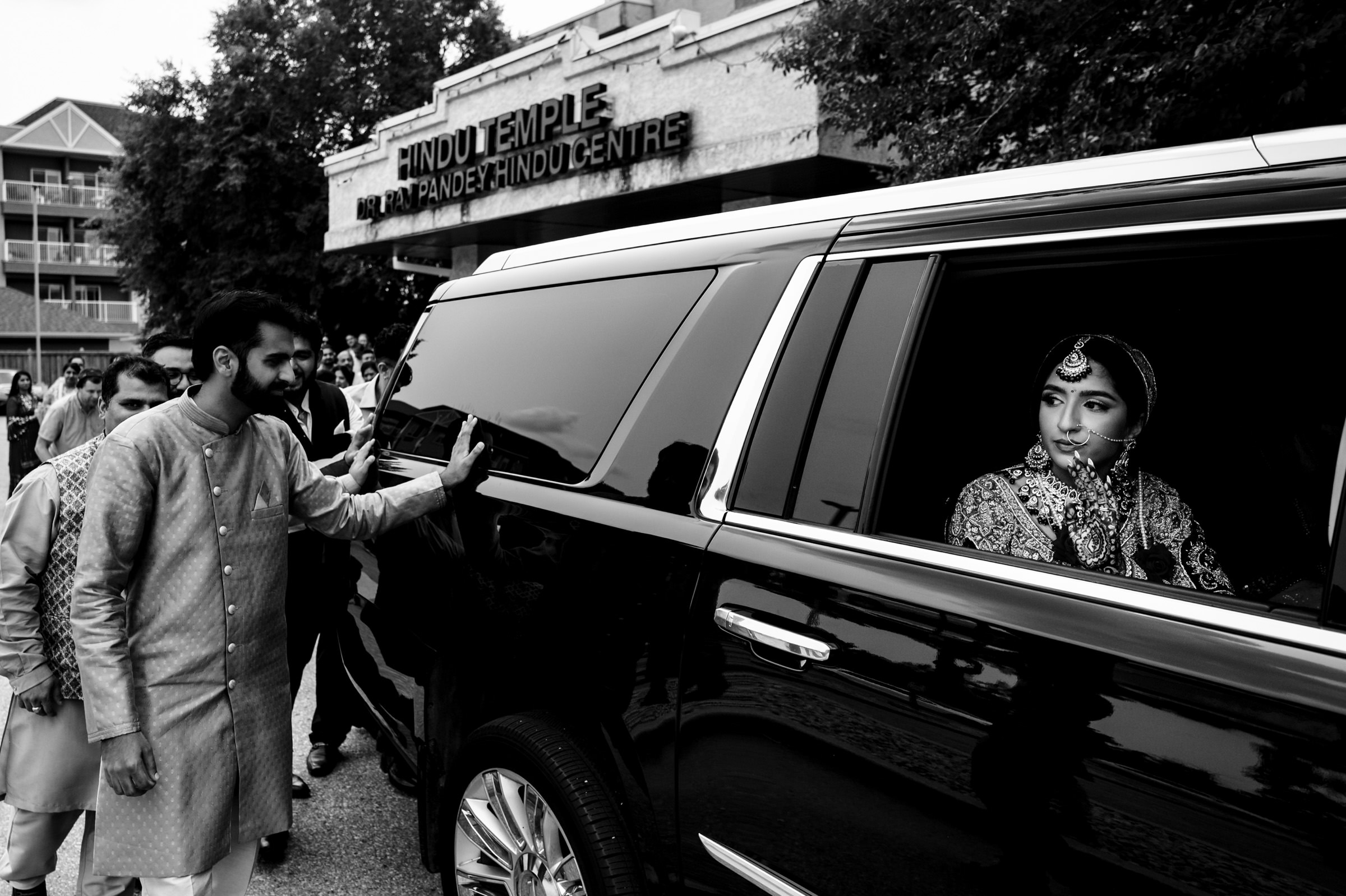 A woman dressed in traditional attire sits inside a car, looking out the window. A man, also in traditional clothing, stands outside the car, touching its door. In the background, a sign reads "Hindu Temple Raj Pandey Hindu Centre.