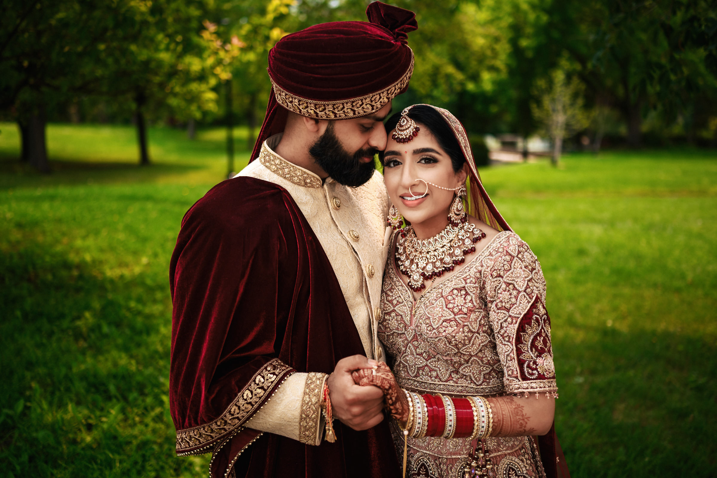 A couple dressed in traditional South Asian wedding attire stands close together. The groom wears a maroon sherwani and turban, while the bride wears a red and gold embroidered lehenga with jewelry. They hold hands and smile against a green, outdoor background.