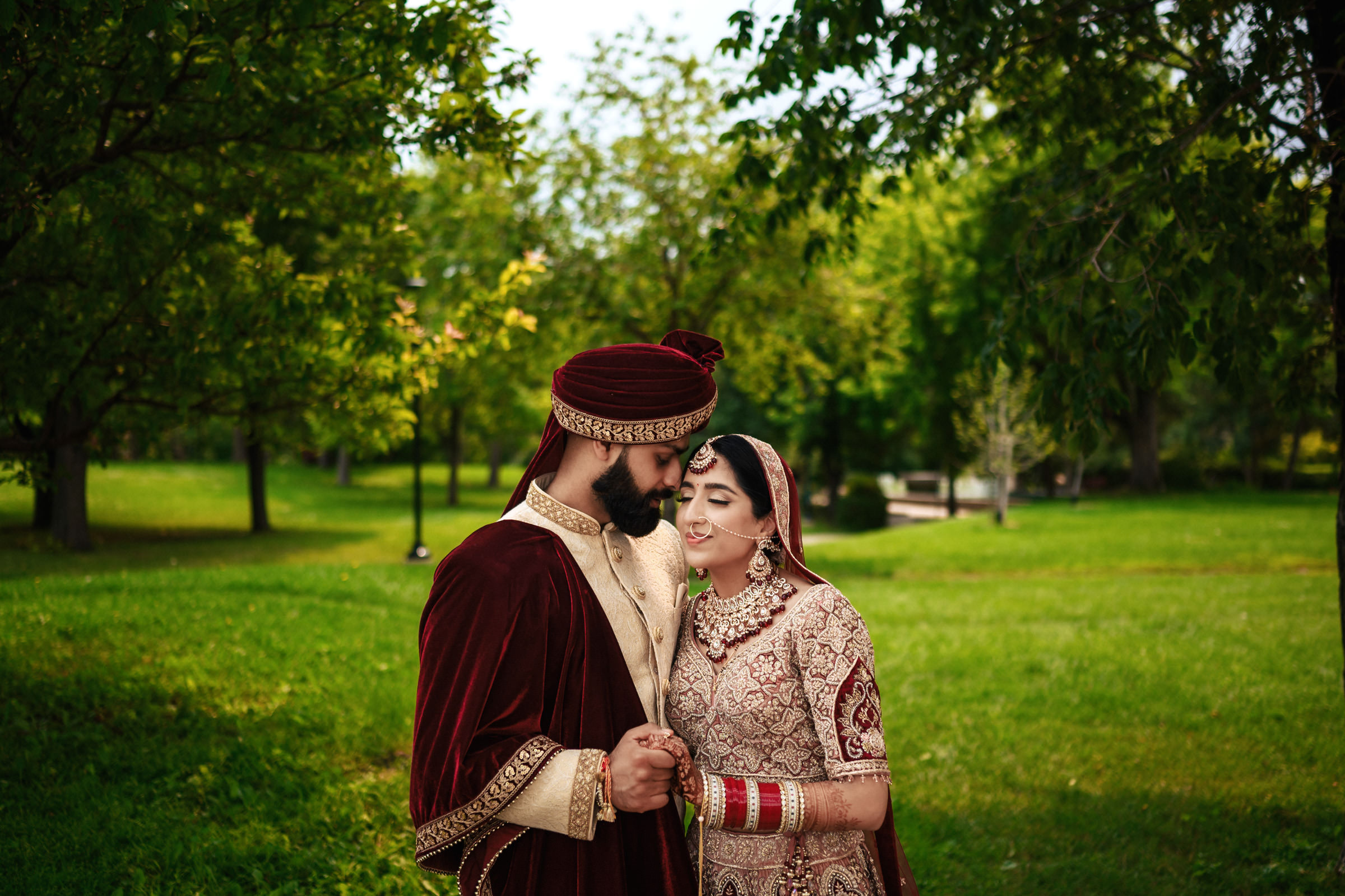 A couple dressed in traditional Indian wedding attire share an intimate moment in a lush green park. The groom, in a maroon and gold sherwani and turban, holds the hand of the bride, who wears an intricately embroidered lehenga with red bangles and jewelry.