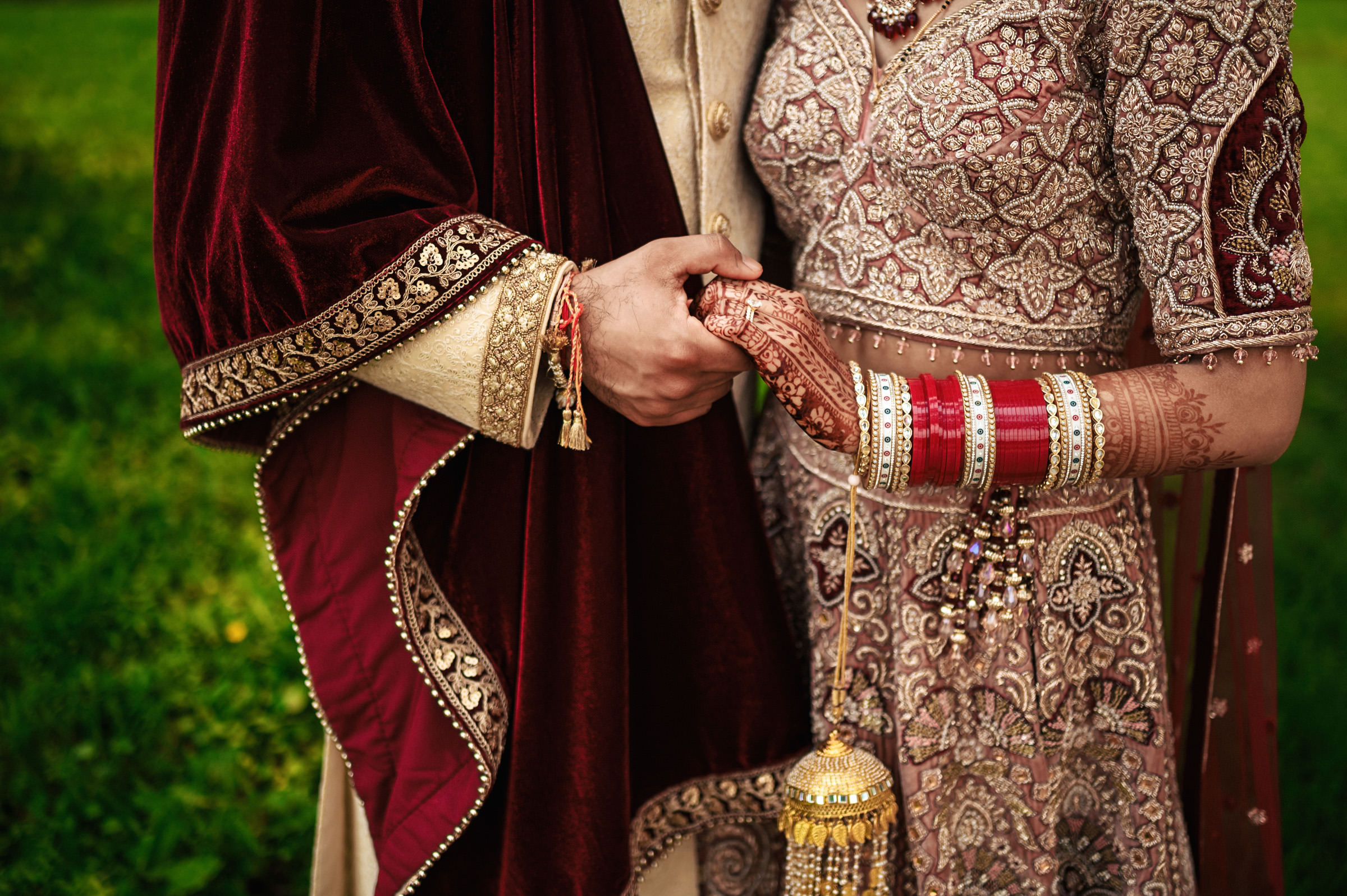 A couple dressed in intricate traditional South Asian wedding attire. The groom wears a beige and maroon sherwani, while the bride dons a detailed lehenga with red and gold bangles. They are holding hands, and the bride's henna-decorated hands are visible.