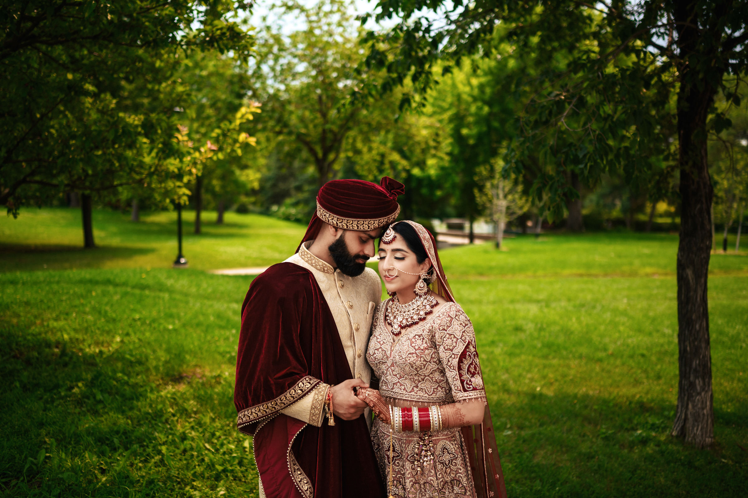 A couple dressed in traditional attire stands closely together on green grass in a park. The man wears a maroon and beige outfit with a turban, while the woman wears a heavily embroidered dress with jewelry and bangles. Both look affectionate and happy.