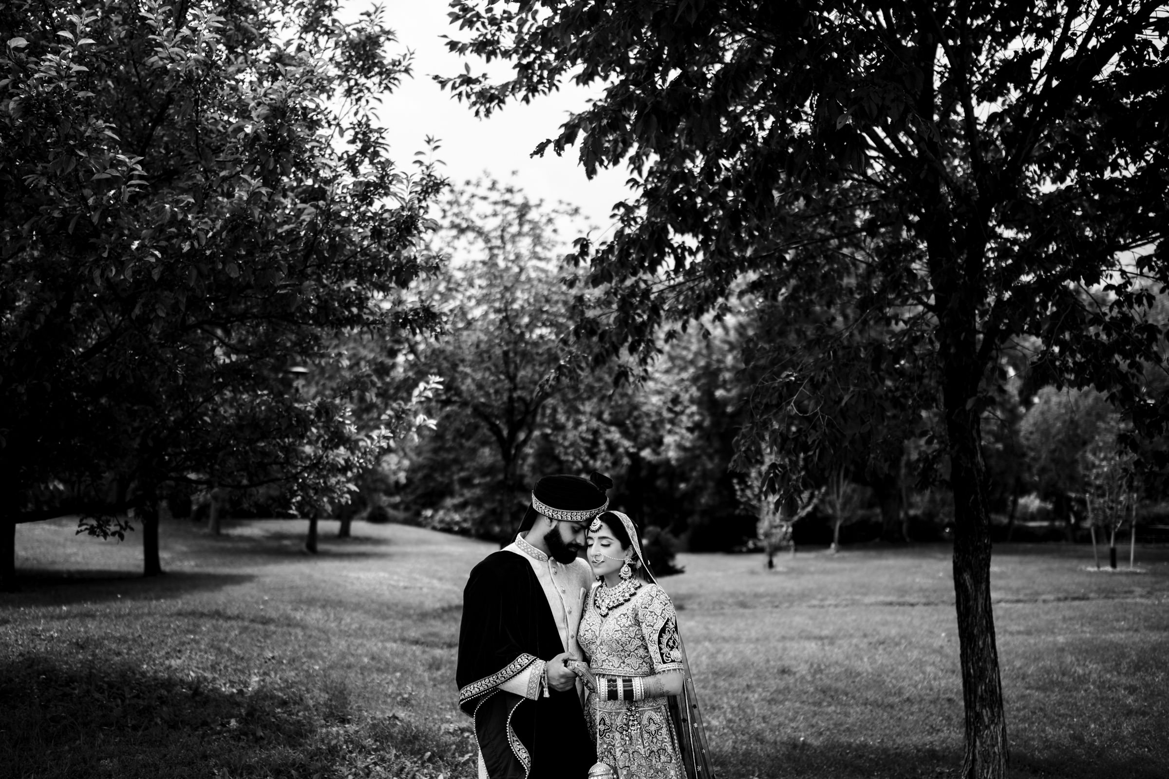 A black-and-white photo of a couple standing close together in a lush, green park with several trees. The man is wearing traditional clothing and a hat, while the woman is dressed in an elaborate gown with a veil. They hold hands and gazing lovingly at each other.