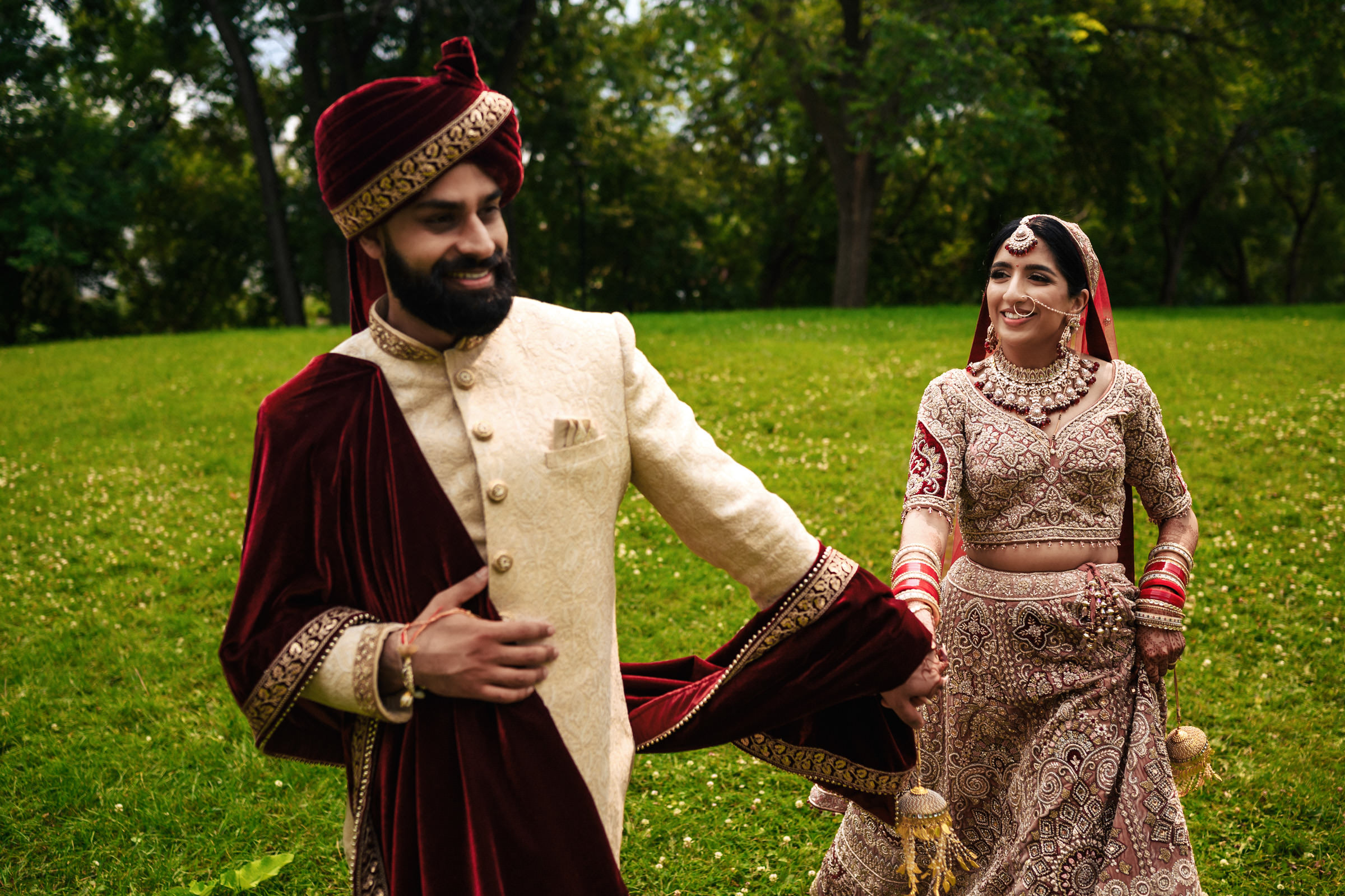 A bride and groom walk hand in hand on a lush green lawn. The groom wears a cream and burgundy sherwani with a matching turban. The bride is dressed in an ornate brown lehenga with red bangles and gold jewelry. Both are smiling and looking at each other affectionately.
