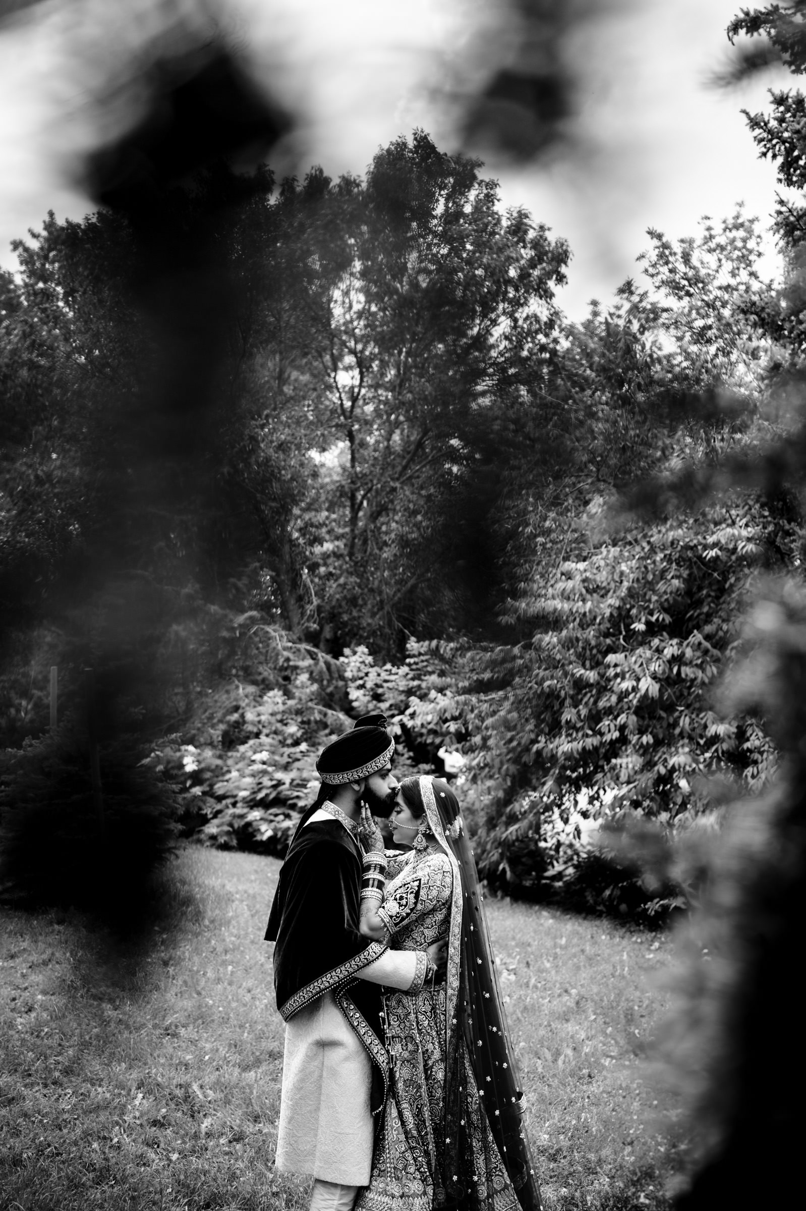 A black and white photo of a couple in traditional attire sharing an intimate moment in a lush garden. The framed shot captures the couple from a distance, surrounded by foliage and trees, creating a romantic and serene ambiance.