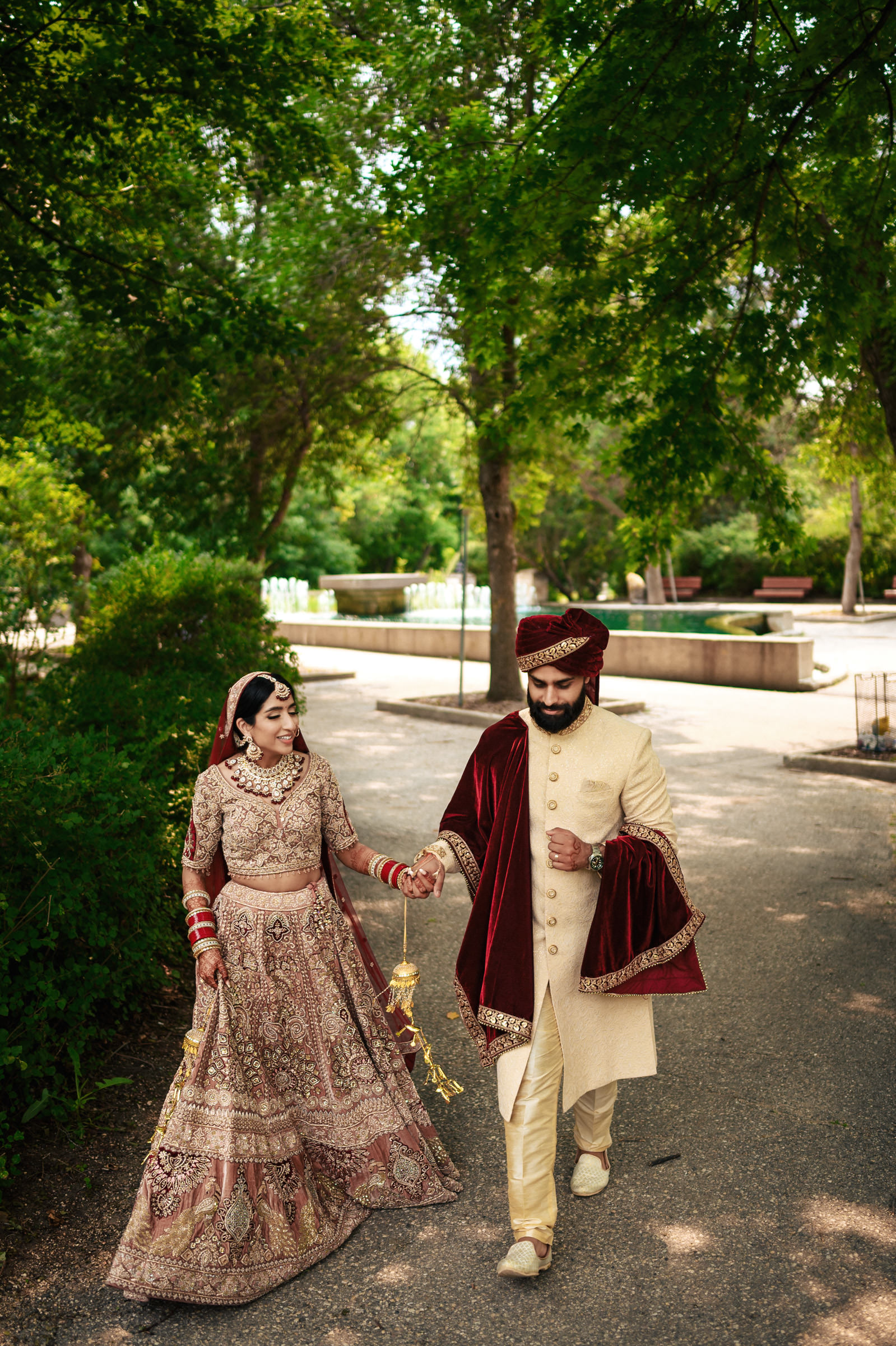 A couple dressed in traditional South Asian wedding attire walks hand in hand down a tree-lined pathway. The bride wears an ornate lehenga, and the groom is dressed in a sherwani. They both smile happily as they walk, surrounded by lush greenery.