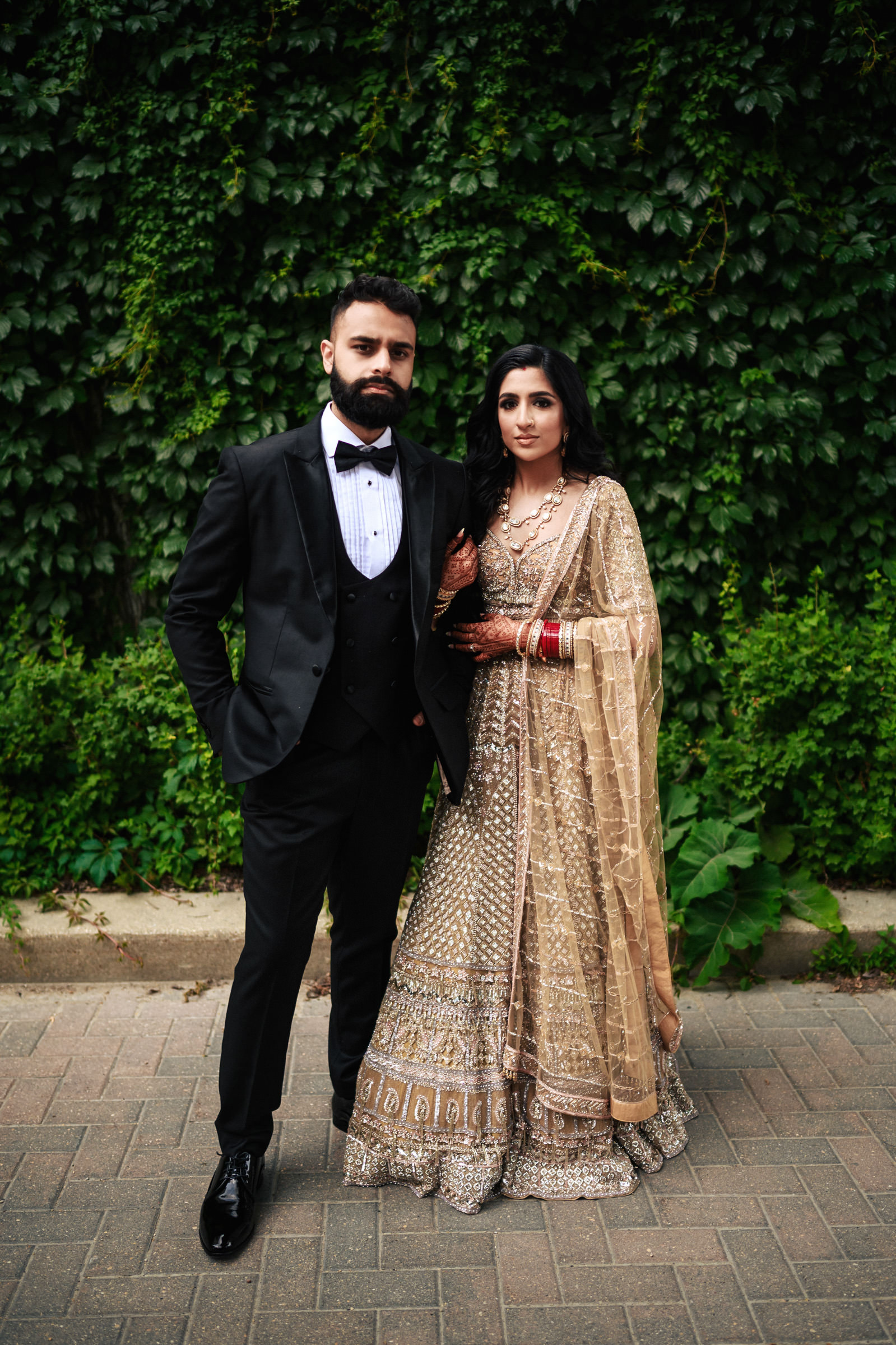 A man and a woman stand side by side in front of a wall covered with green foliage. The man is dressed in a black tuxedo with a bow tie, and the woman is wearing a traditional decorated gown with red bangles. Both have dark hair and serious expressions.