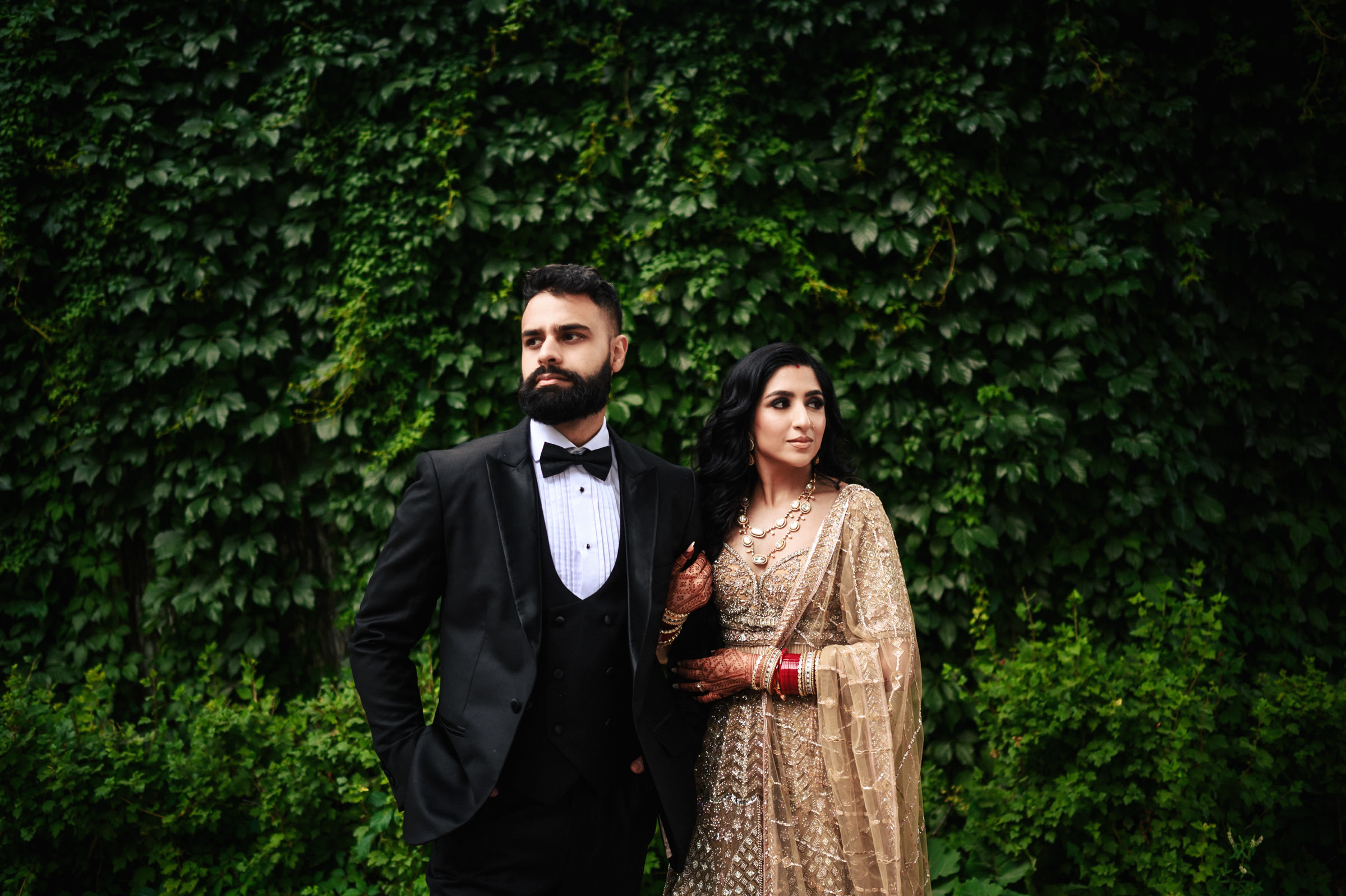 A man and woman stand side by side in formal attire against a lush, green foliage backdrop. The man wears a black tuxedo with a bow tie, while the woman is dressed in an ornate beige saree with henna on her hands. Both look off into the distance.