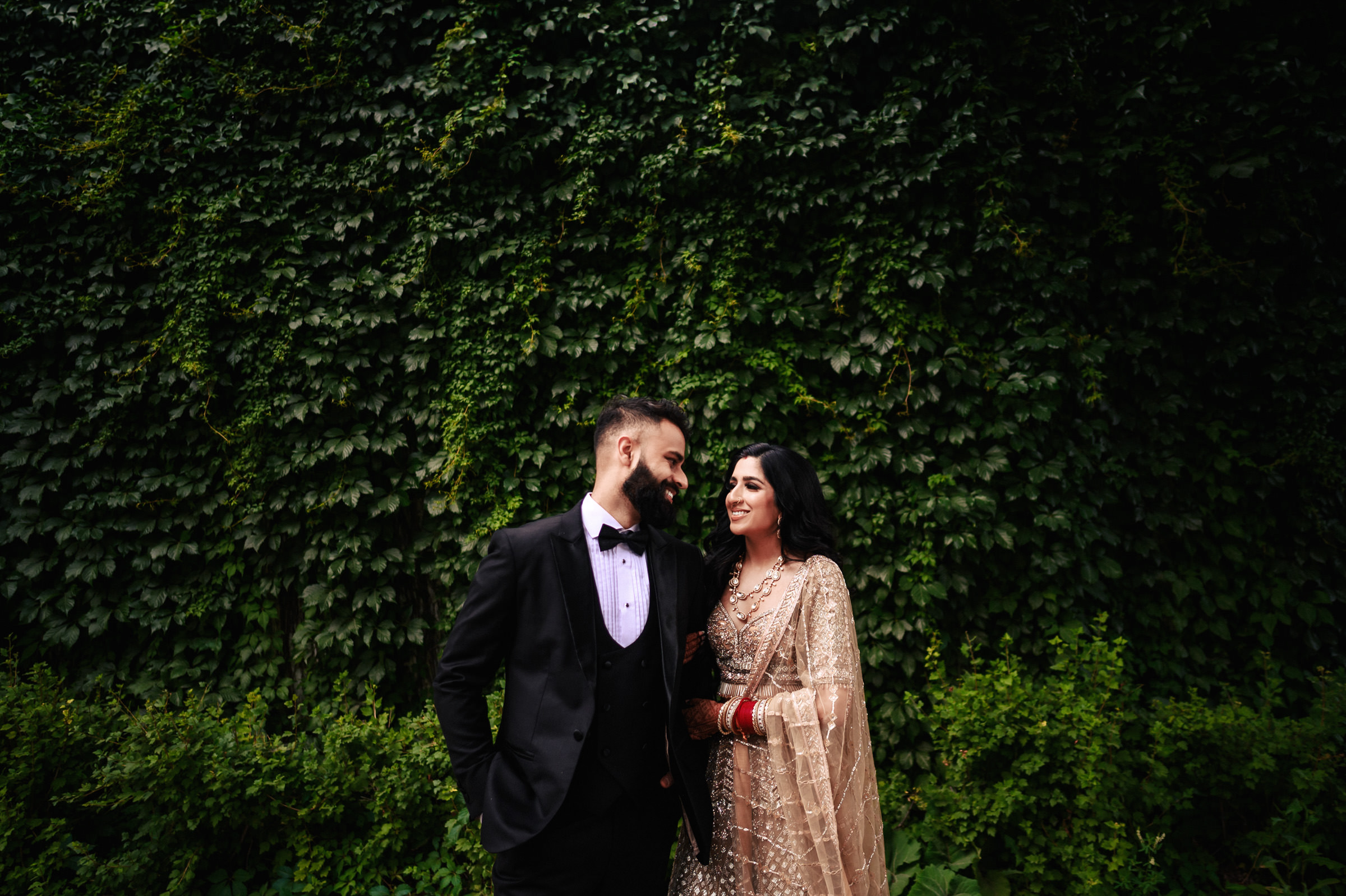 A couple stands closely in front of a lush, green ivy-covered wall. The man wears a black suit with a bow tie, while the woman is dressed in an ornate, gold traditional outfit. Both are smiling and looking at each other warmly.