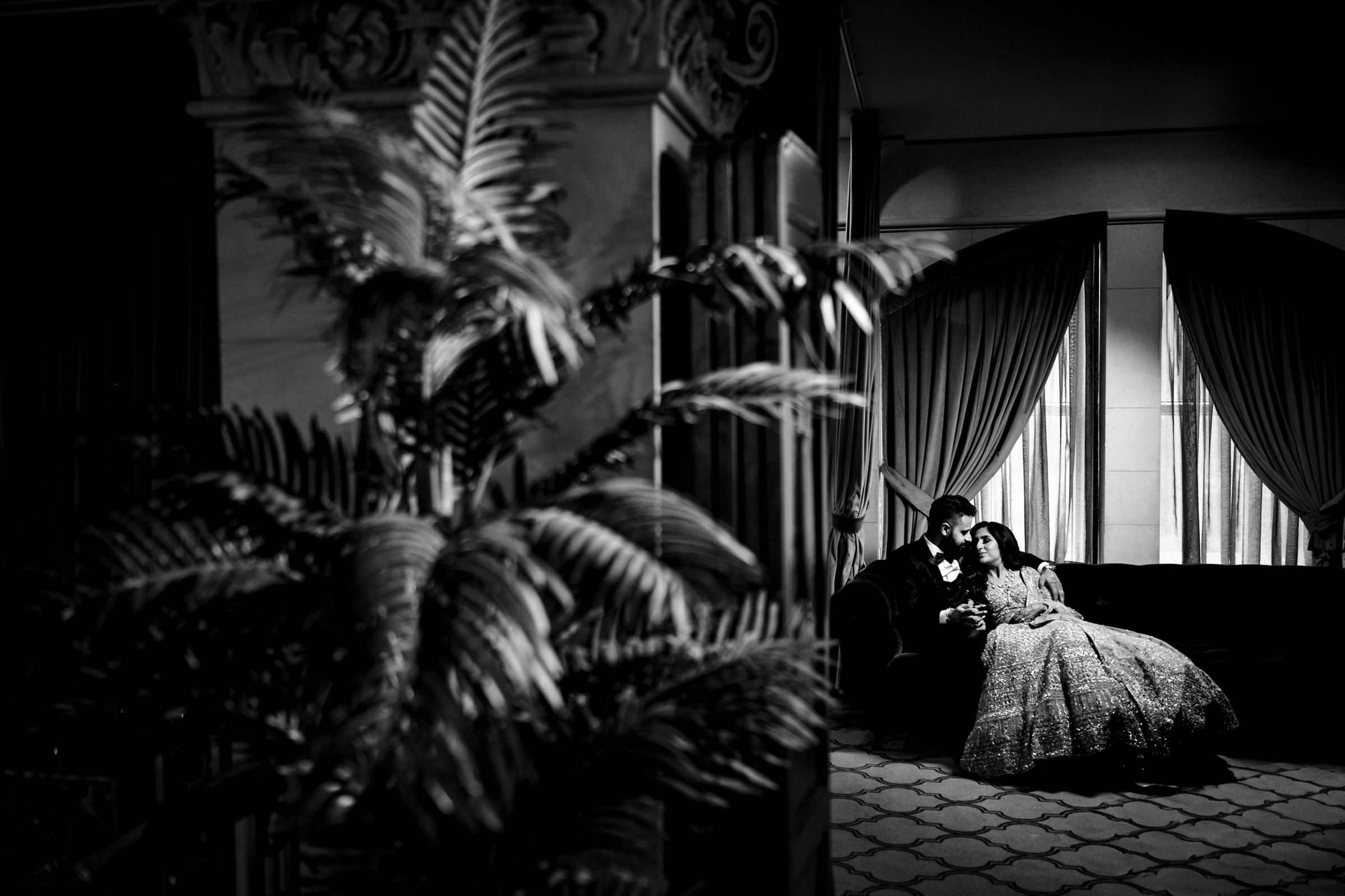 A black-and-white photograph depicting a couple sitting closely on a plush sofa in a dimly lit room. The woman is resting her head on the man's shoulder. Large, elegant curtains adorn the windows in the background, and a tall leafy plant is in the foreground on the left.